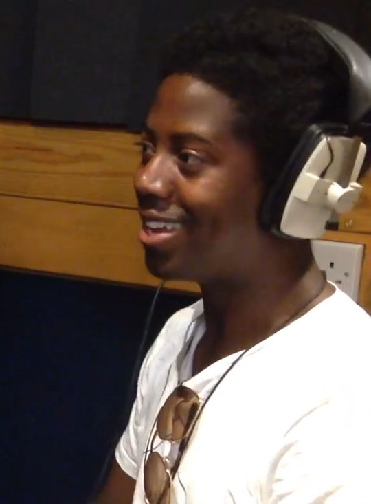 Moses Boyd laughs mid-sentence in a recording booth, wearing over-ear headphones and a white T-shirt with sunglasses hooked at the collar.