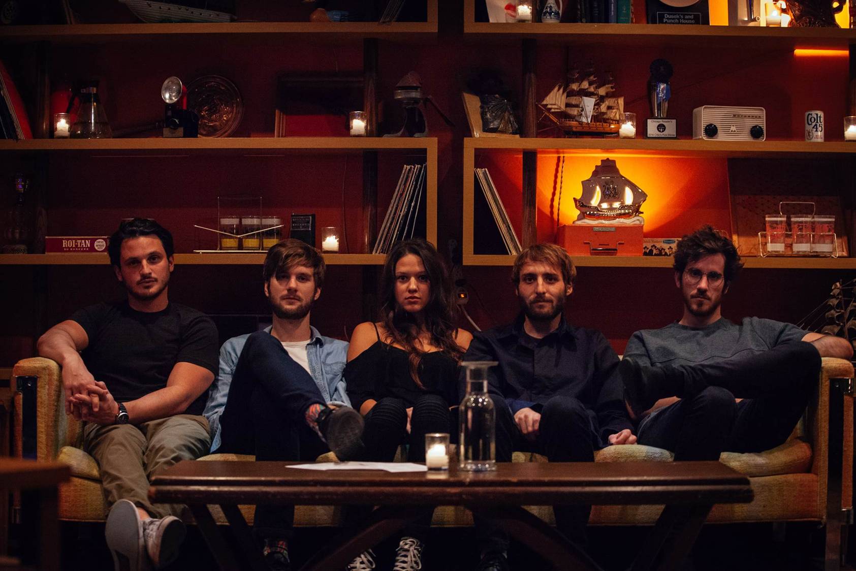 The five members of Mt. Joy sit side by side on a low couch in a candlelit bar, shelves of bottles, records and a model ship behind them.