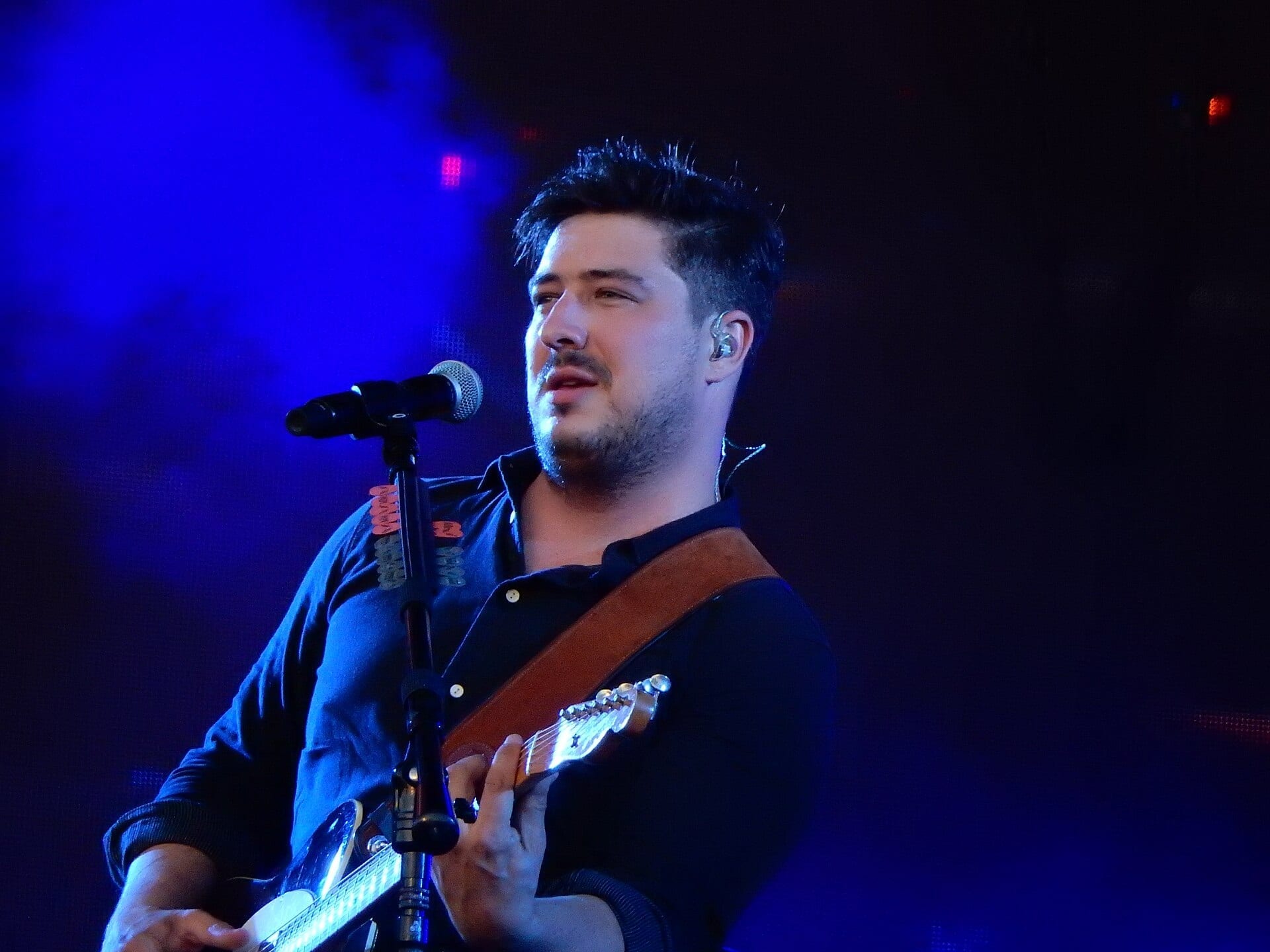 Marcus Mumford of Mumford & Sons sings into a microphone while playing guitar on stage, lit by blue stage light with an in-ear monitor visible.