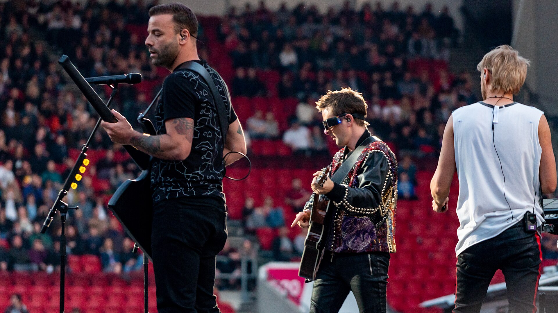 Three members of Muse on a stadium stage, a bassist at a mic stand on the left, Matt Bellamy in sunglasses and a studded jacket playing acoustic guitar in the middle and the drummer standing with his back to the camera on the right