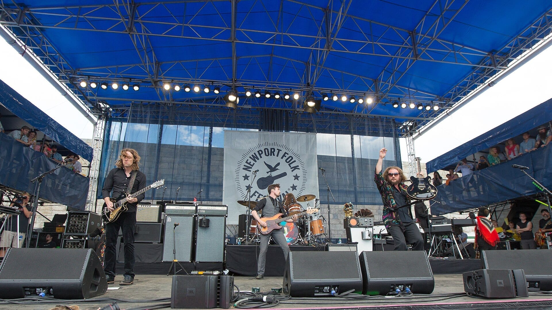 My Morning Jacket performs on an outdoor stage under a blue canopy, with frontman Jim James raising a fist beside a banner reading Newport Folk Festival.