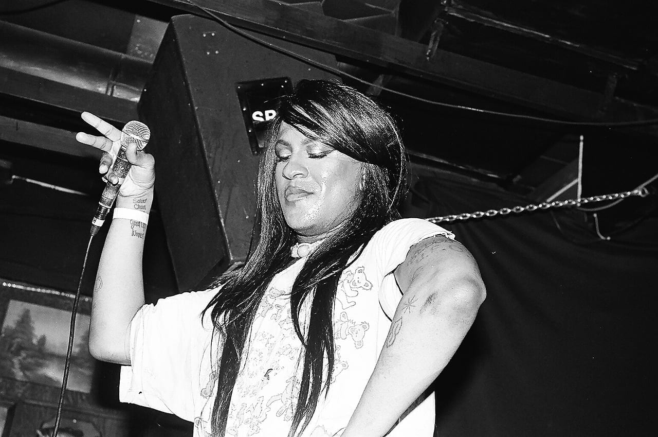 Mykki Blanco performs on stage in black and white, holding a microphone with eyes closed and long hair falling across the face.