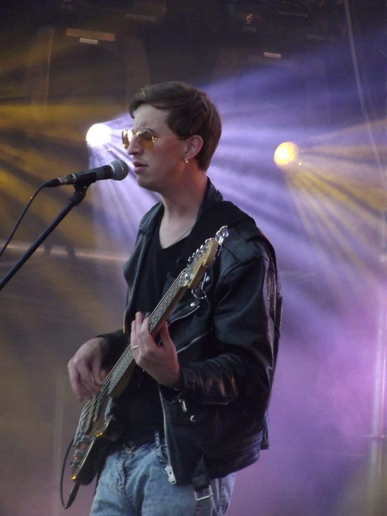 A bassist in tinted glasses and a black leather jacket steps up to a microphone under purple and yellow stage lights.