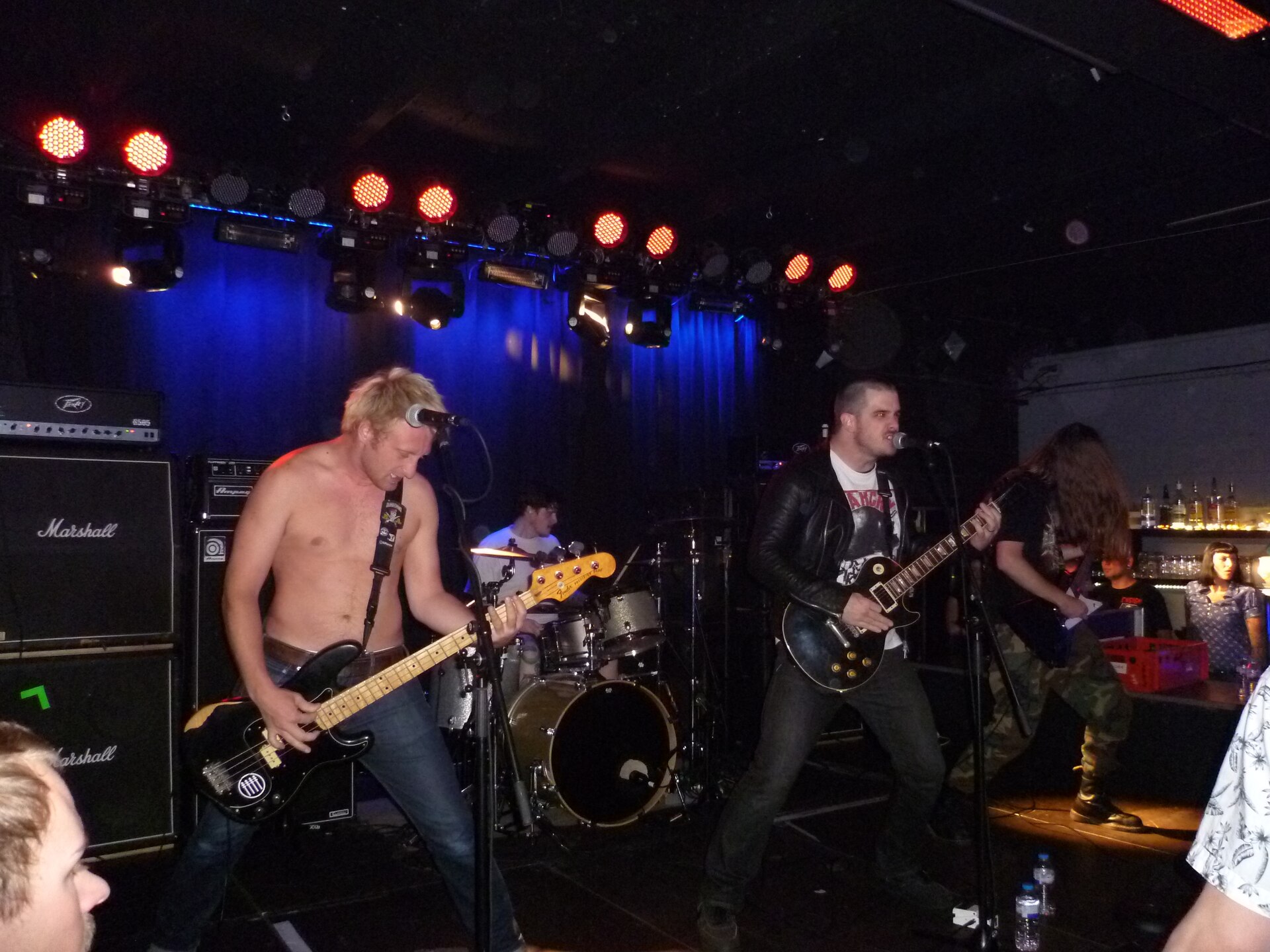 A cramped club stage with Nails mid-set, the bassist bent over his instrument on the left and the guitarist shouting into a microphone on the right, Marshall cabinets stacked beside them.
