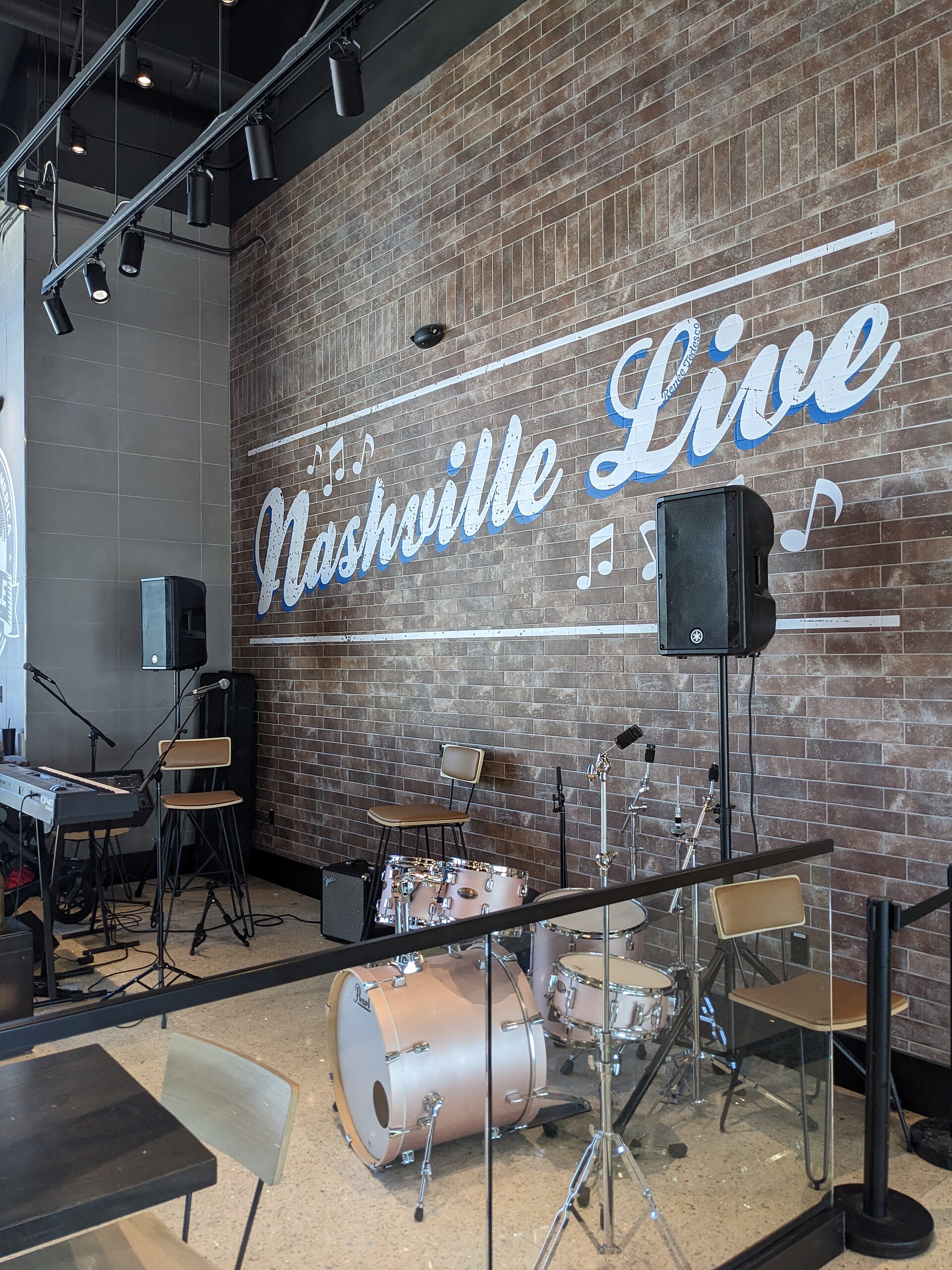 A small live-music stage inside Nashville International Airport, with a drum kit, keyboard and PA speakers set against a brick wall painted Nashville Live.