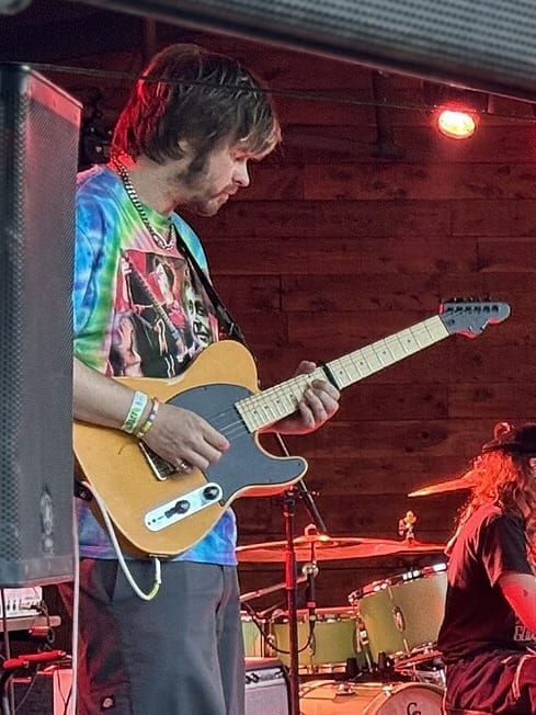 Nate Amos playing an electric guitar on an outdoor stage, wearing a tie-dye t-shirt, with a drummer visible behind him.