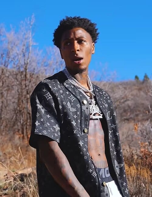 NBA YoungBoy stands outdoors in front of dry brush and a clear blue sky, wearing an open patterned shirt and layered chains.