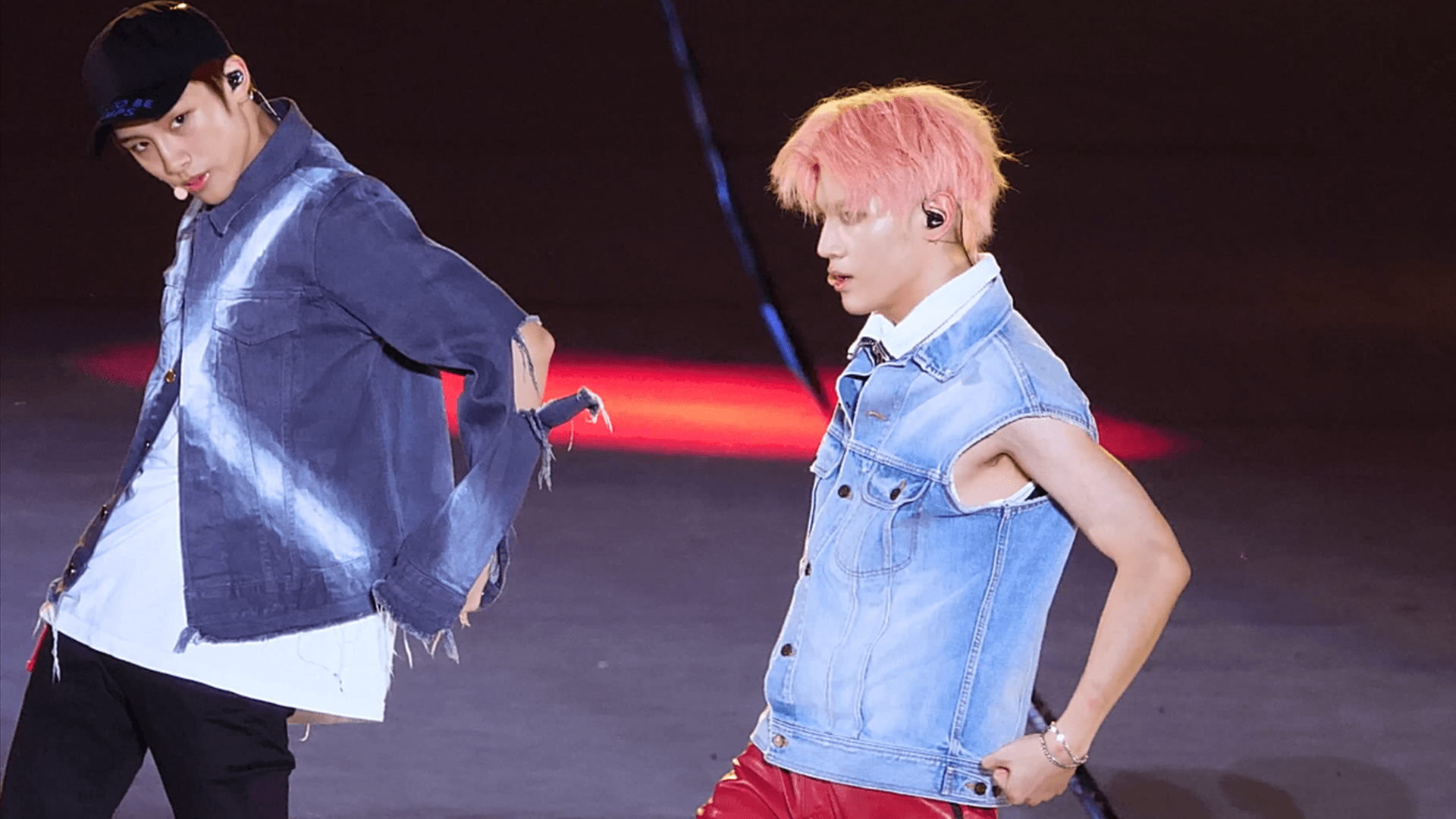 Two NCT 127 members hold a dance pose mid-routine on a dark arena stage, one in a cap and cut denim jacket, the other in a sleeveless denim vest with pink hair.