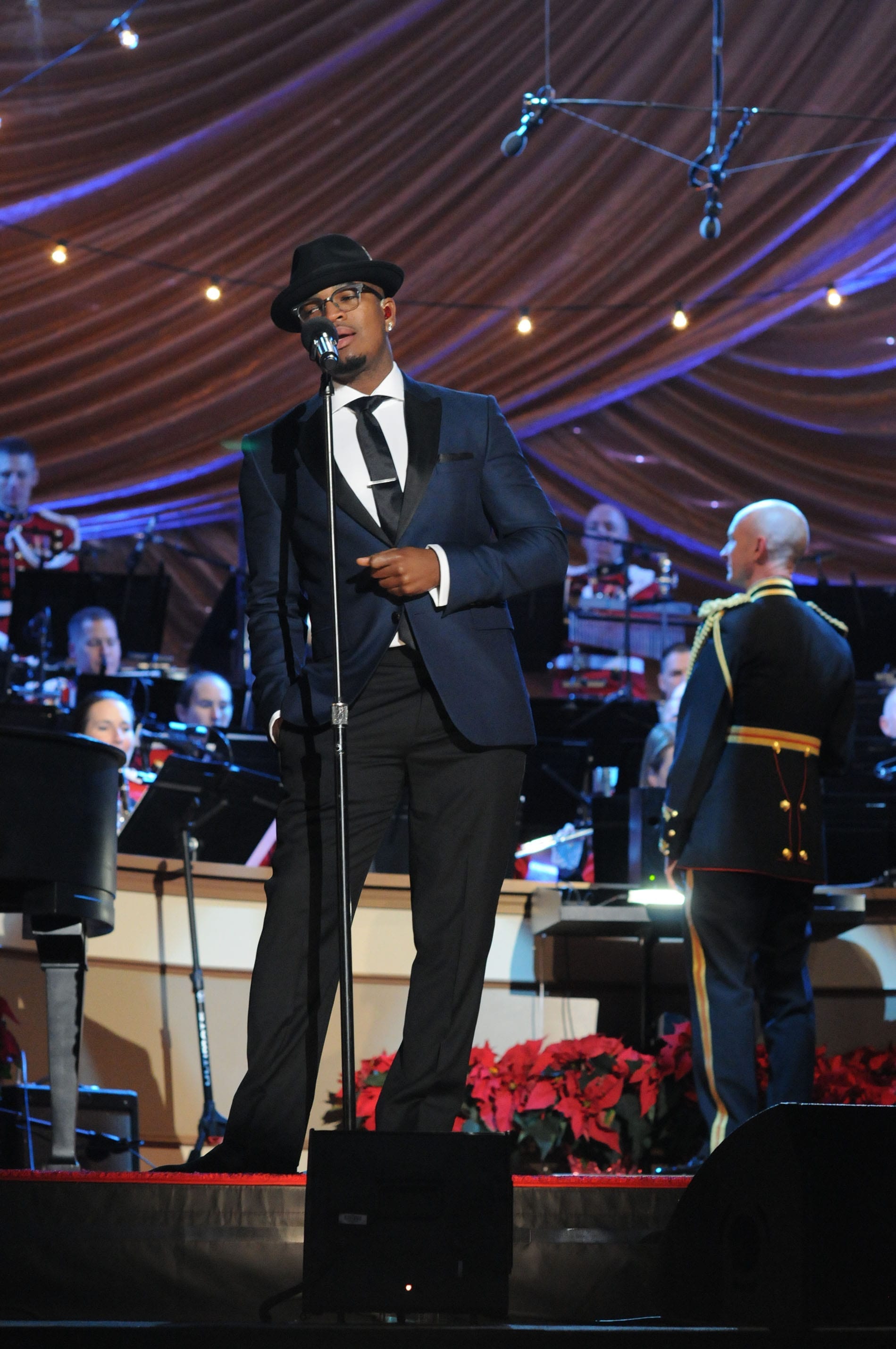 Ne-Yo sings at a microphone stand in a dark suit and black hat, an orchestra and rows of poinsettias behind him.