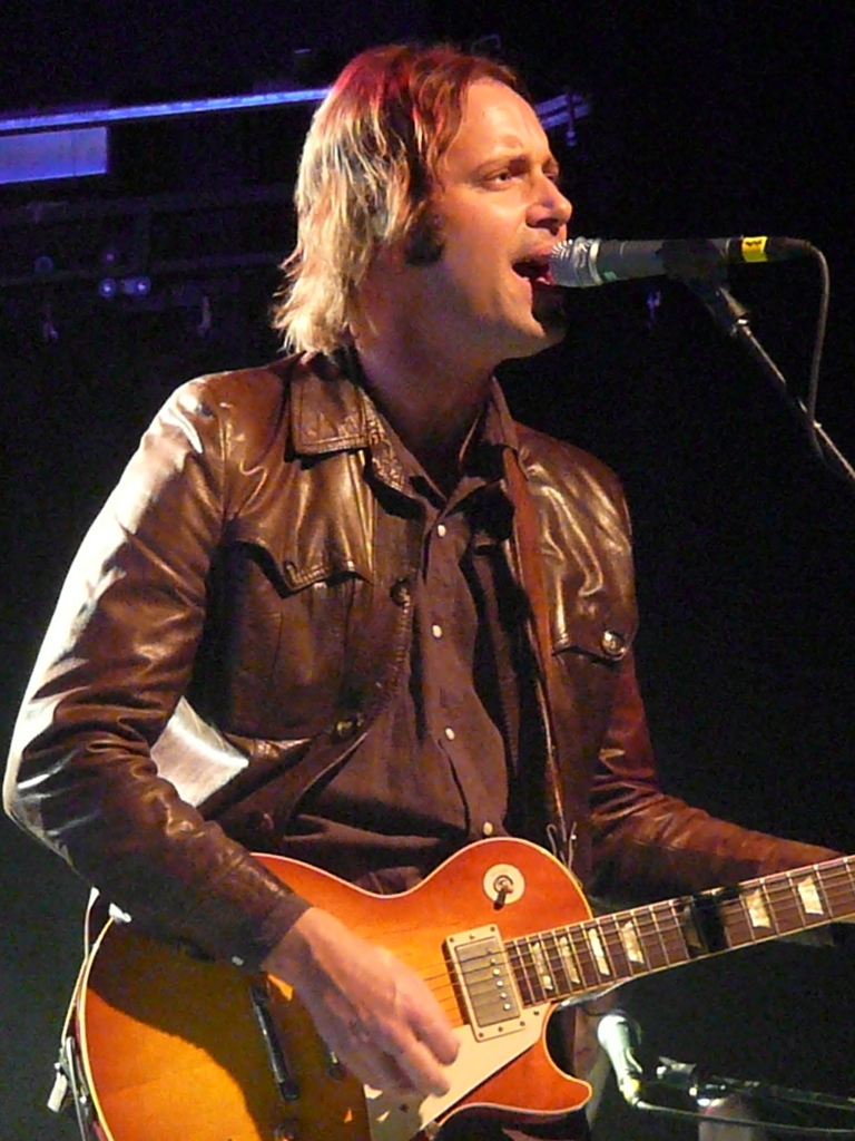 A long-haired man in a brown leather jacket sings into a microphone while playing a sunburst Les Paul on a dark stage