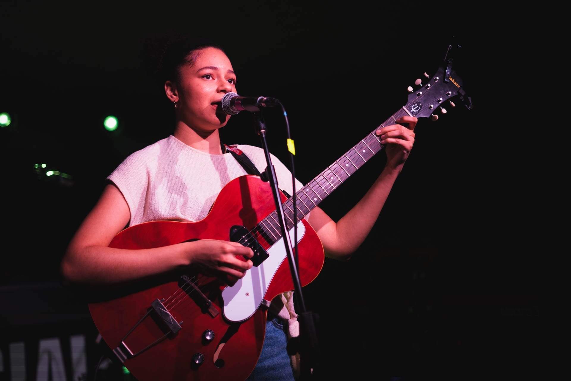 Nectar Woode sings into a microphone on a dark stage while playing a red hollow-body electric guitar, wearing a light patterned top and small hoop earrings, with green stage lights behind her.