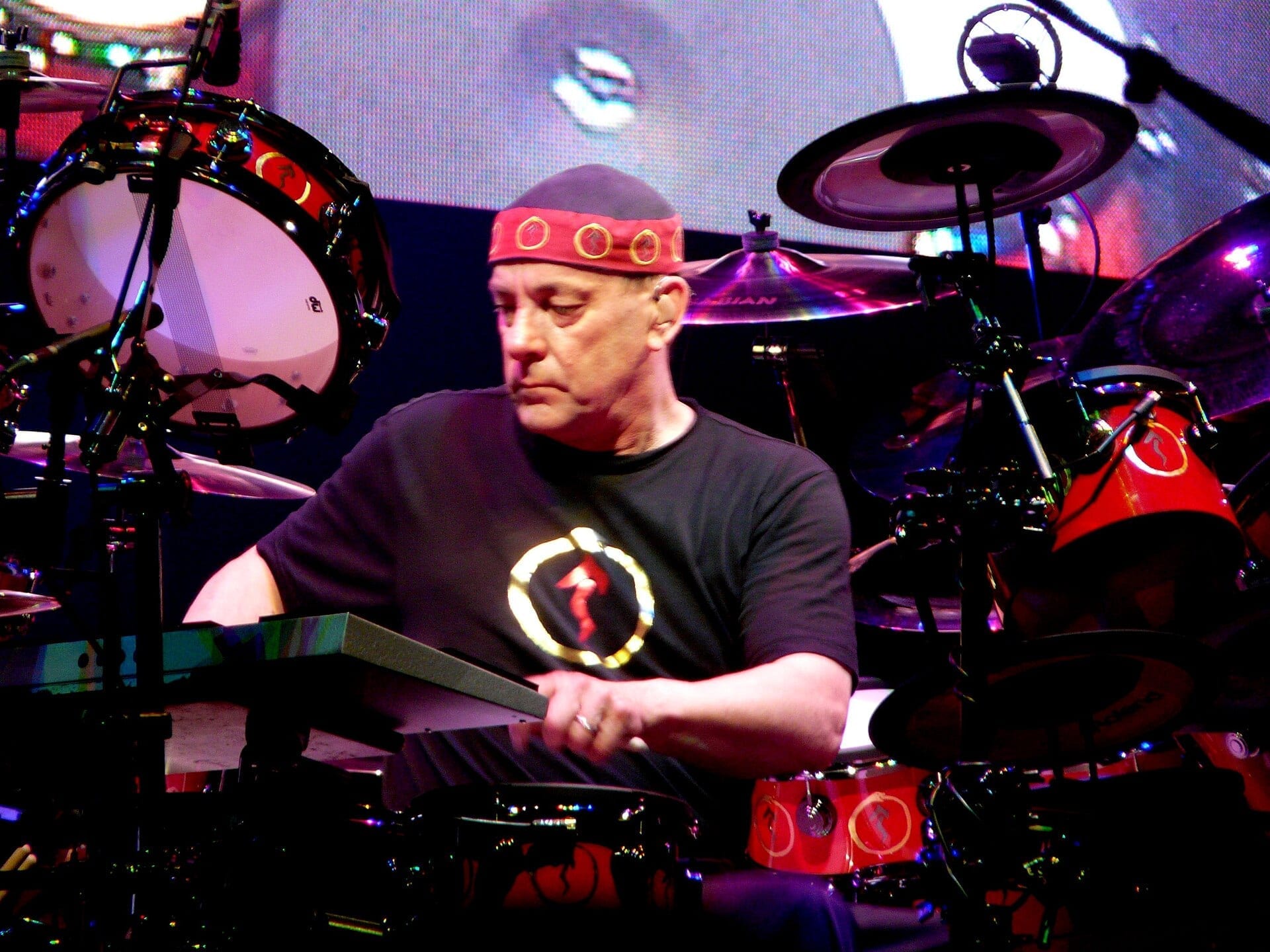 Neil Peart looks down at his snare mid-fill, in a red patterned headband and a black T-shirt, ringed by red and black drums and cymbals.