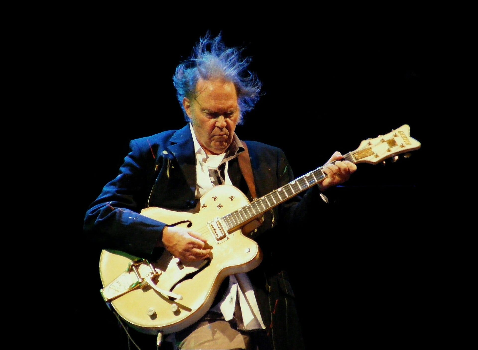 Neil Young, his hair blown back, plays a cream-colored electric guitar on a dark stage, wearing a navy blazer over a white shirt.