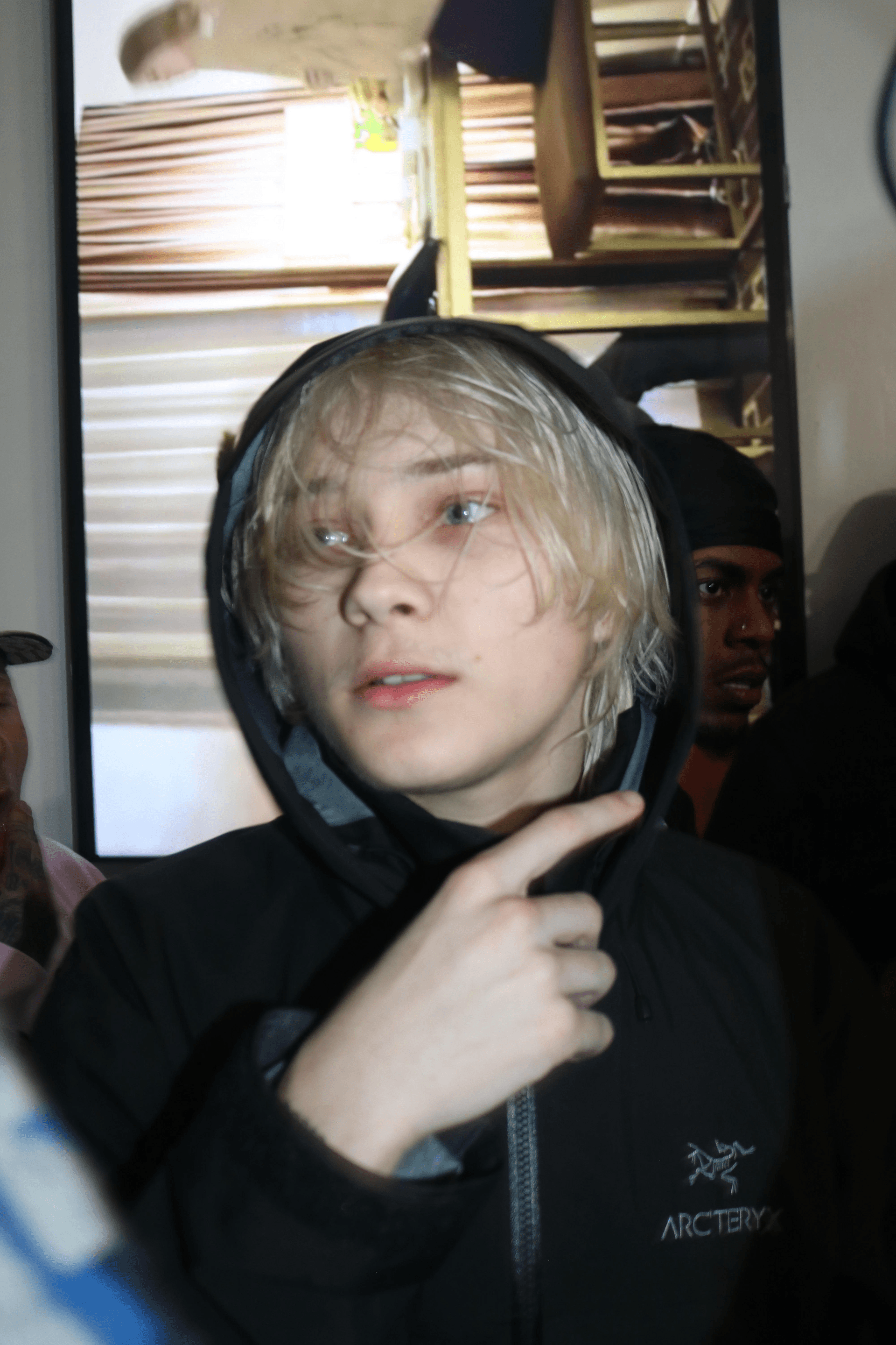 Nettspend, a young rapper with bleach-blond hair, wearing a black hooded jacket and looking off to the side indoors.