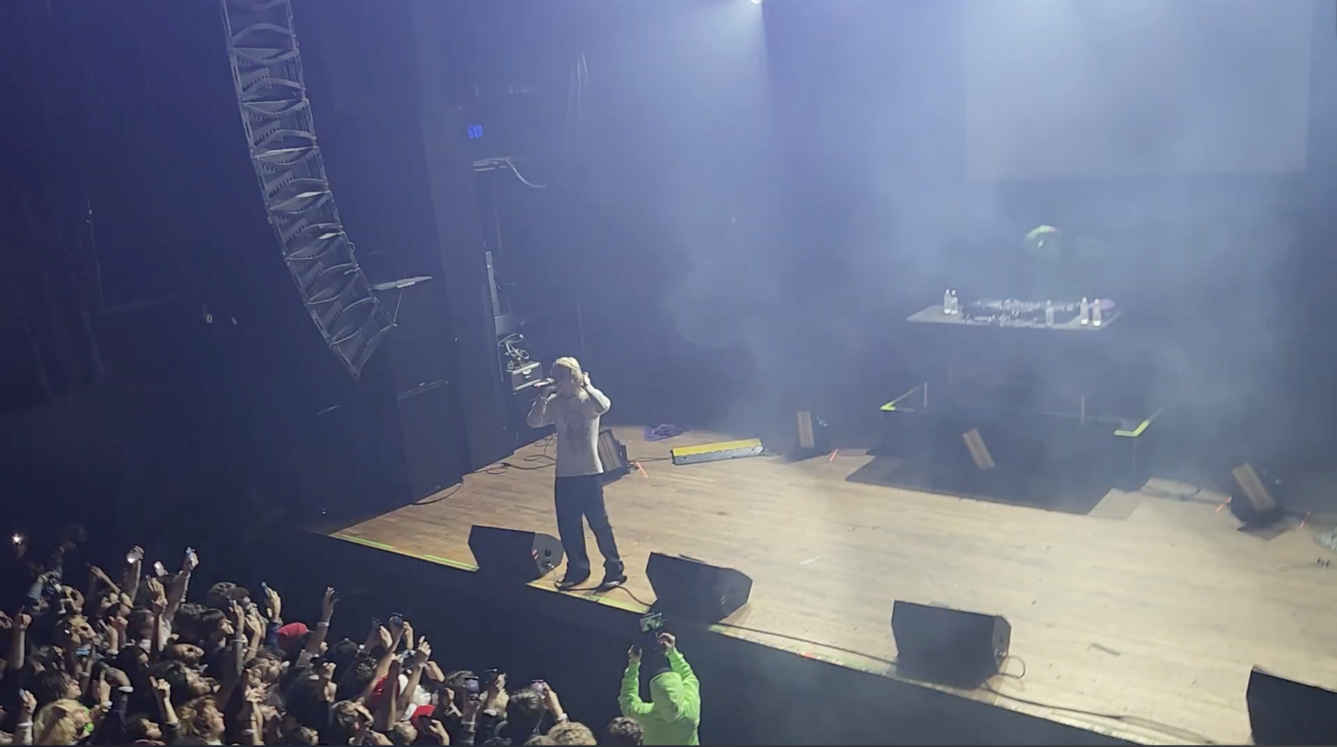 A lone performer stands at the front of a smoky theatre stage with a microphone raised, a crowd of lifted phones below the stage lip.