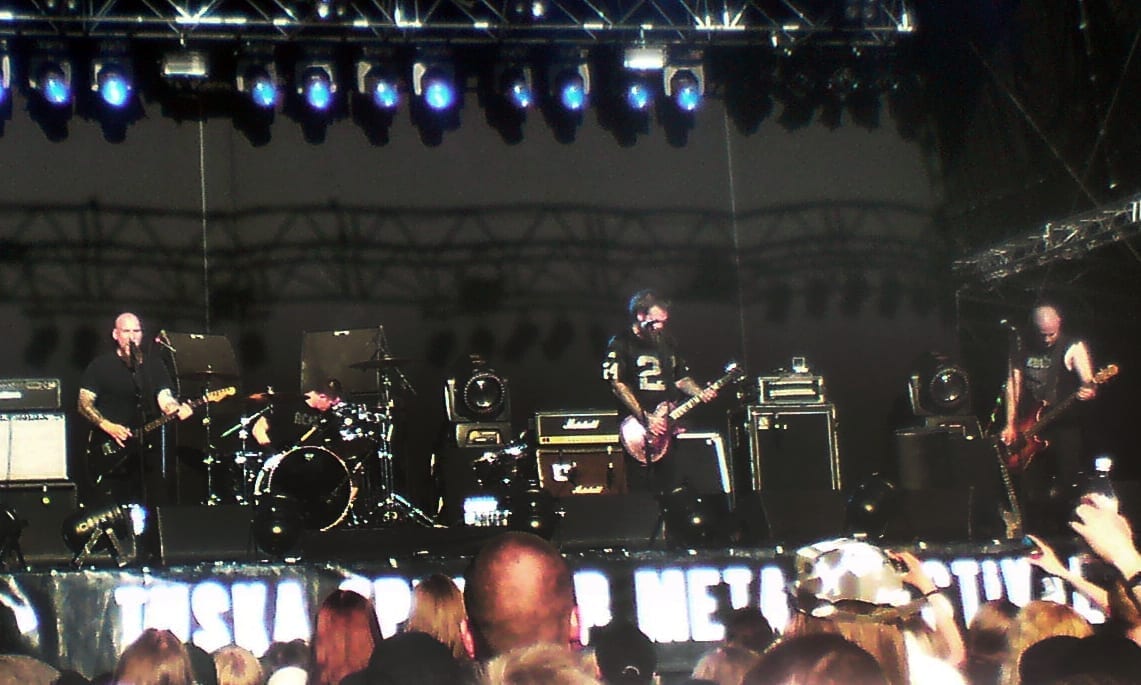 Four members of Neurosis perform on an outdoor festival stage at night under blue lighting, with a crowd in front and a Tuska festival banner visible below the stage.