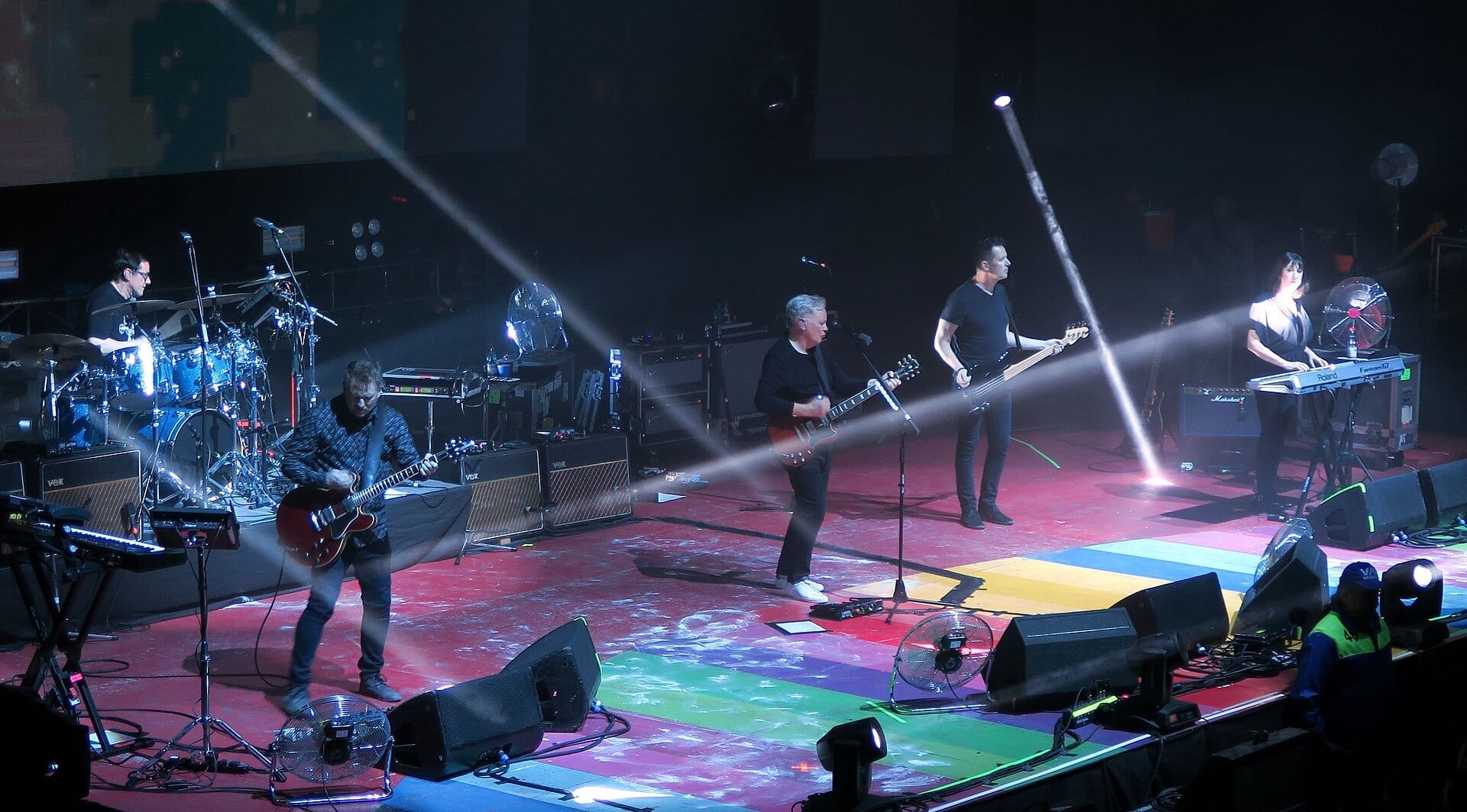 The five members of New Order perform on a rainbow-lit stage with drums, guitars, bass and keyboards, crossed by two bright stage-light beams.