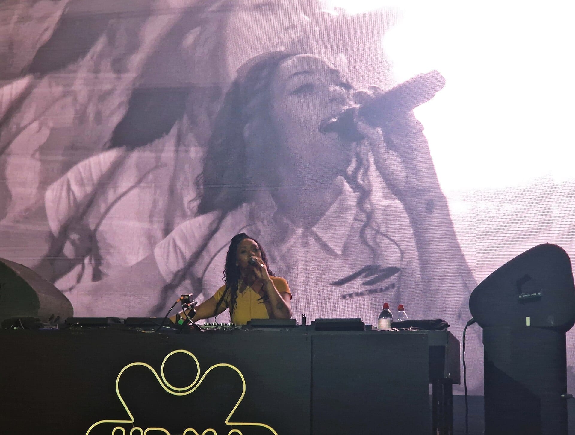 Nia Archives, in a yellow top, sings into a handheld microphone at a DJ booth marked Speakerbox, with a large video screen behind her showing a close-up of the same moment.