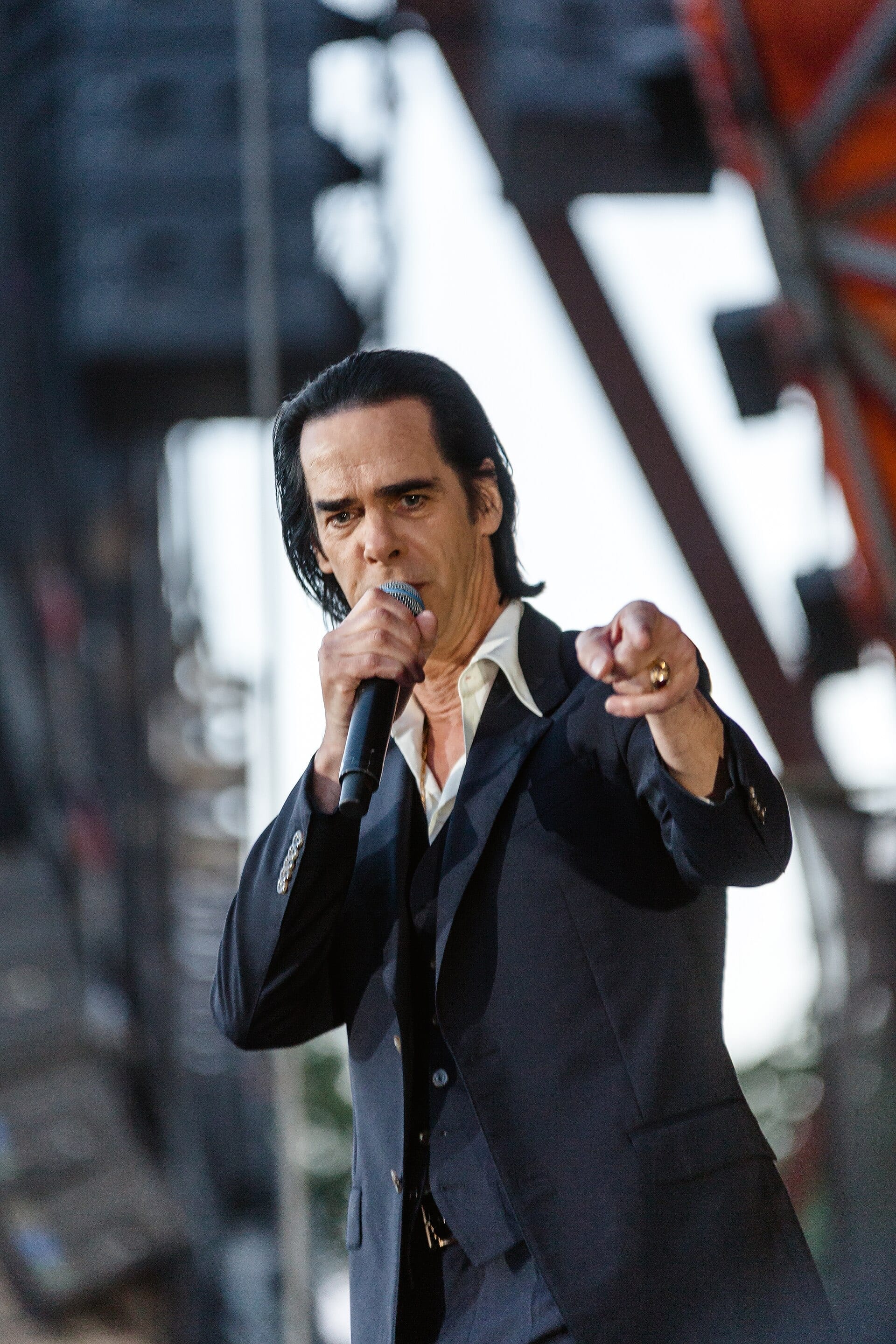Nick Cave sings into a microphone and points toward the camera on an outdoor festival stage, wearing a dark suit.
