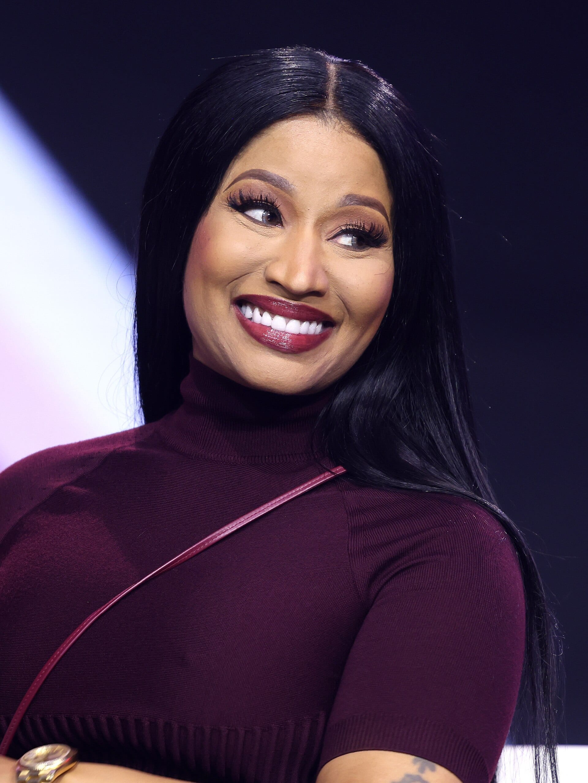 Nicki Minaj smiles and looks off to the side, wearing a dark maroon turtleneck top with long straight black hair.