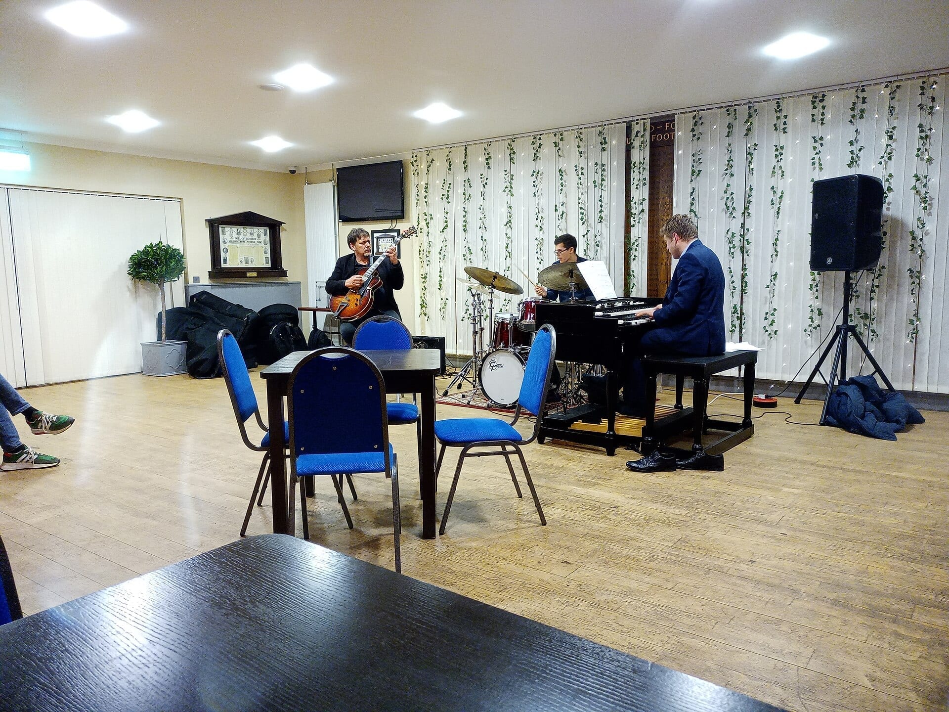 Nigel Price plays a hollow-body jazz guitar in a small community hall, standing next to a drummer and a musician seated at a Hammond organ.