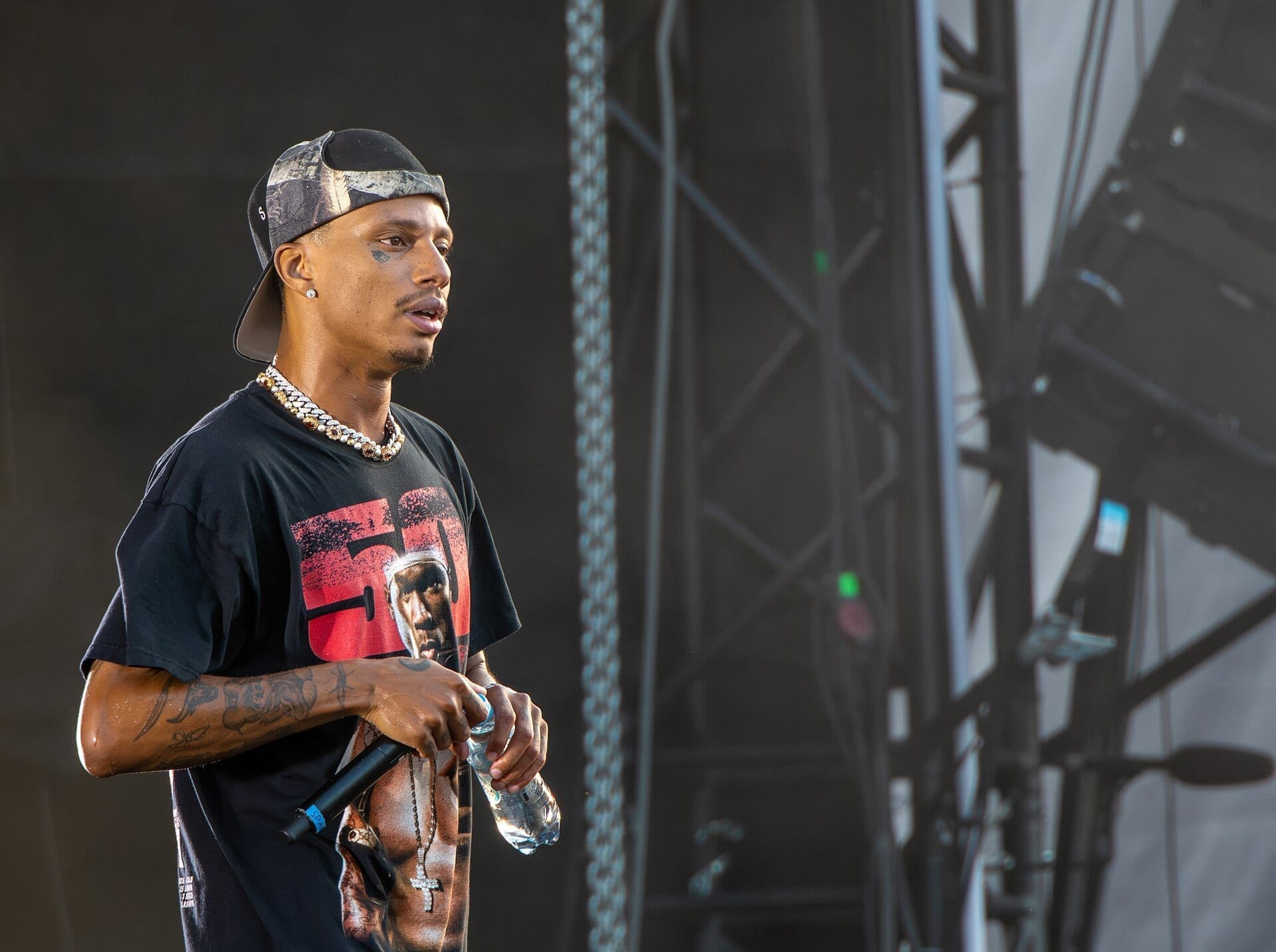 Night Lovell stands in profile on a festival stage in a backwards cap and a graphic tee, an iced chain at his neck, a microphone in one hand and a water bottle in the other.