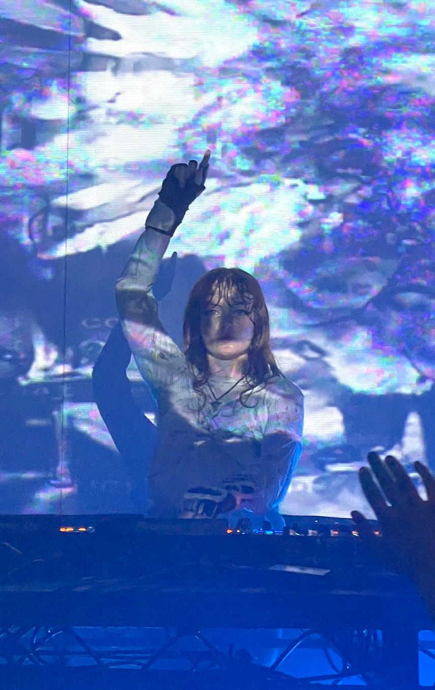 Ninajirachi behind a DJ controller with one gloved hand raised, lit blue, a large glitching projection filling the screen behind her