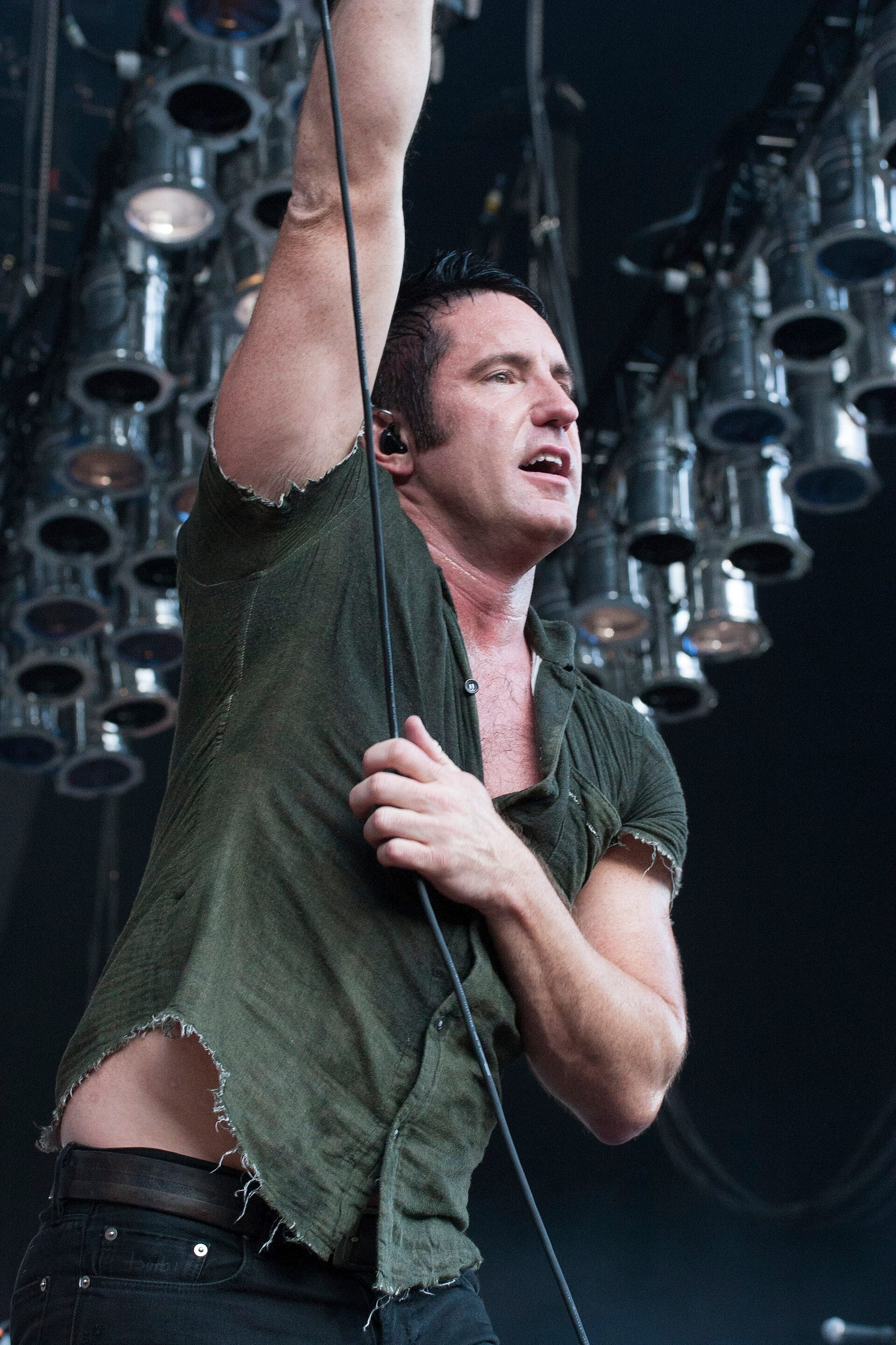 Trent Reznor of Nine Inch Nails sings into a microphone with one arm raised overhead, wearing a torn olive-green shirt on a darkened stage lined with lighting rigs.