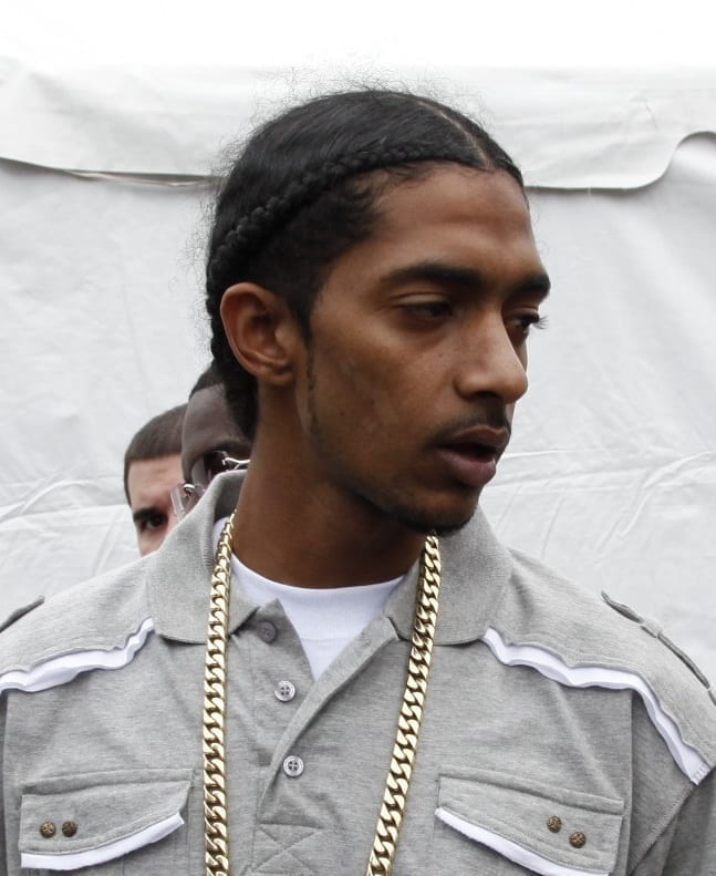 Nipsey Hussle, wearing braided hair and a gold chain over a grey collared shirt, outdoors in 2009.