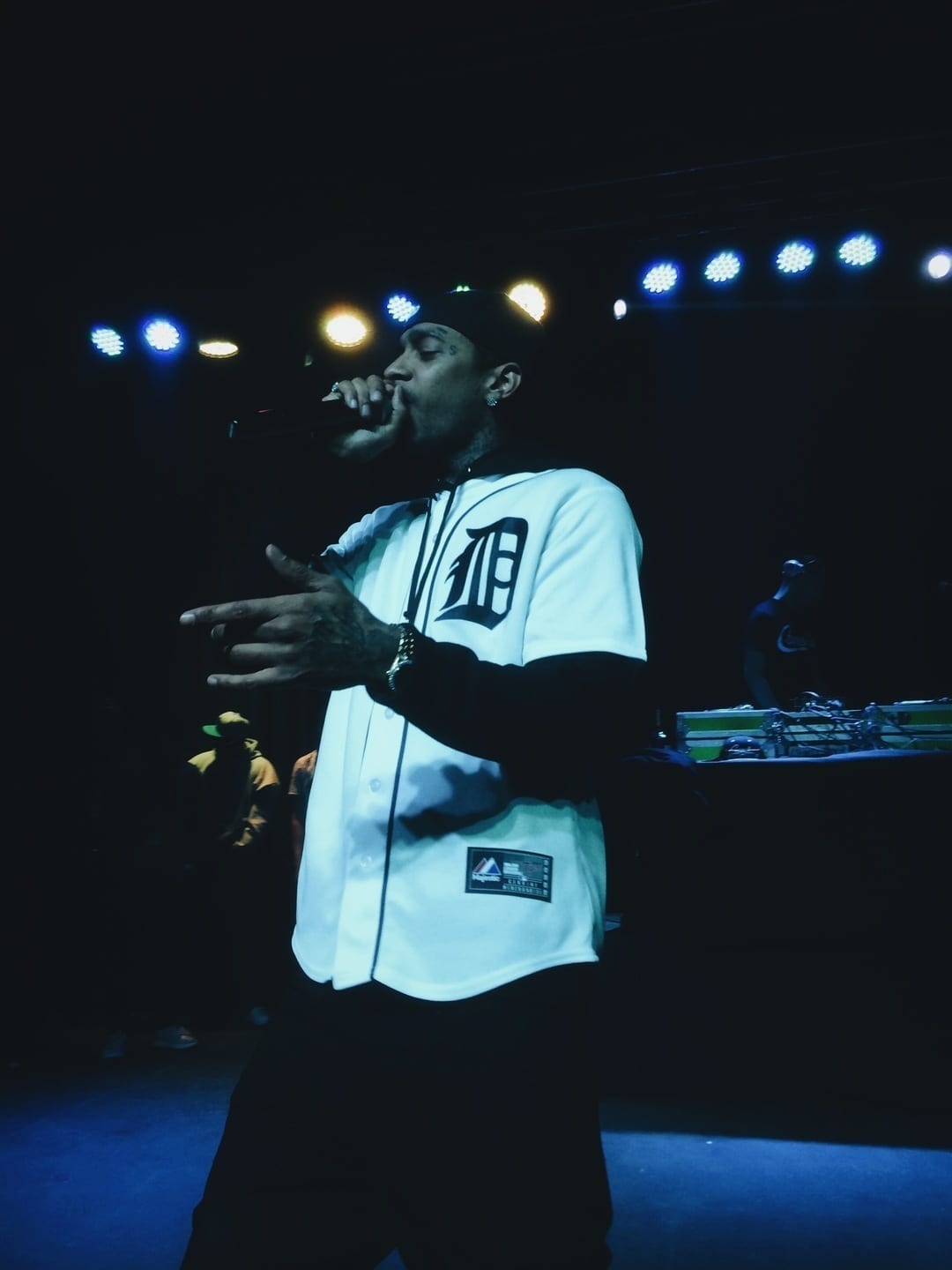 Nipsey Hussle performs on stage into a microphone, wearing a white and black baseball-style jersey with a large letter D, in front of colorful stage lights.