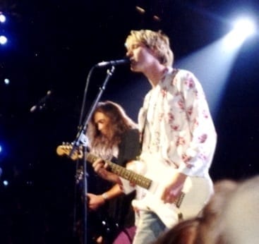 Kurt Cobain sings into a microphone while playing guitar in a floral shirt, with Krist Novoselic playing bass beside him, lit from behind on a dark stage.