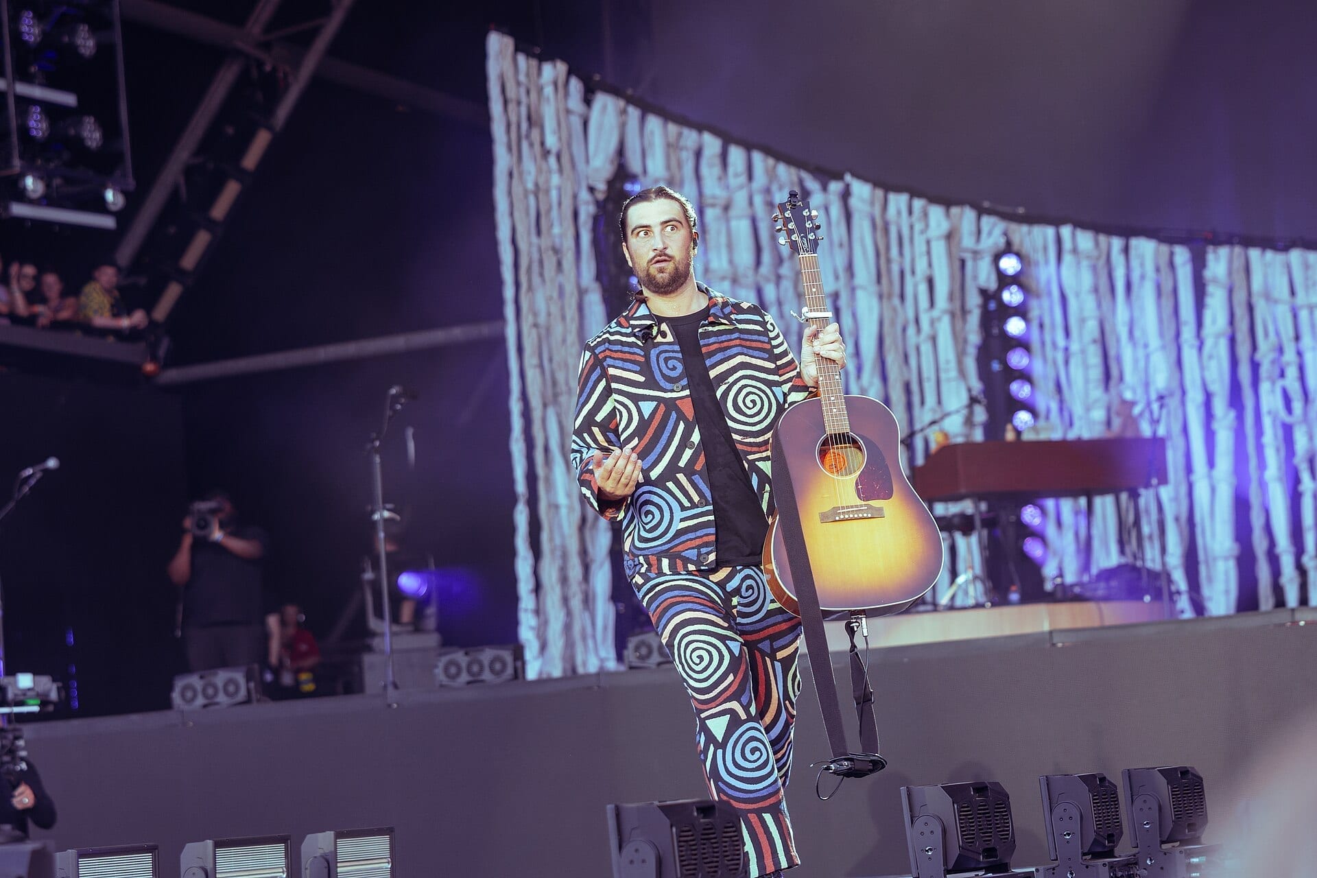 Noah Kahan performs on an outdoor festival stage in a bold spiral-patterned outfit, holding an acoustic guitar and glancing to the side, with a lit backdrop resembling birch trees behind him.