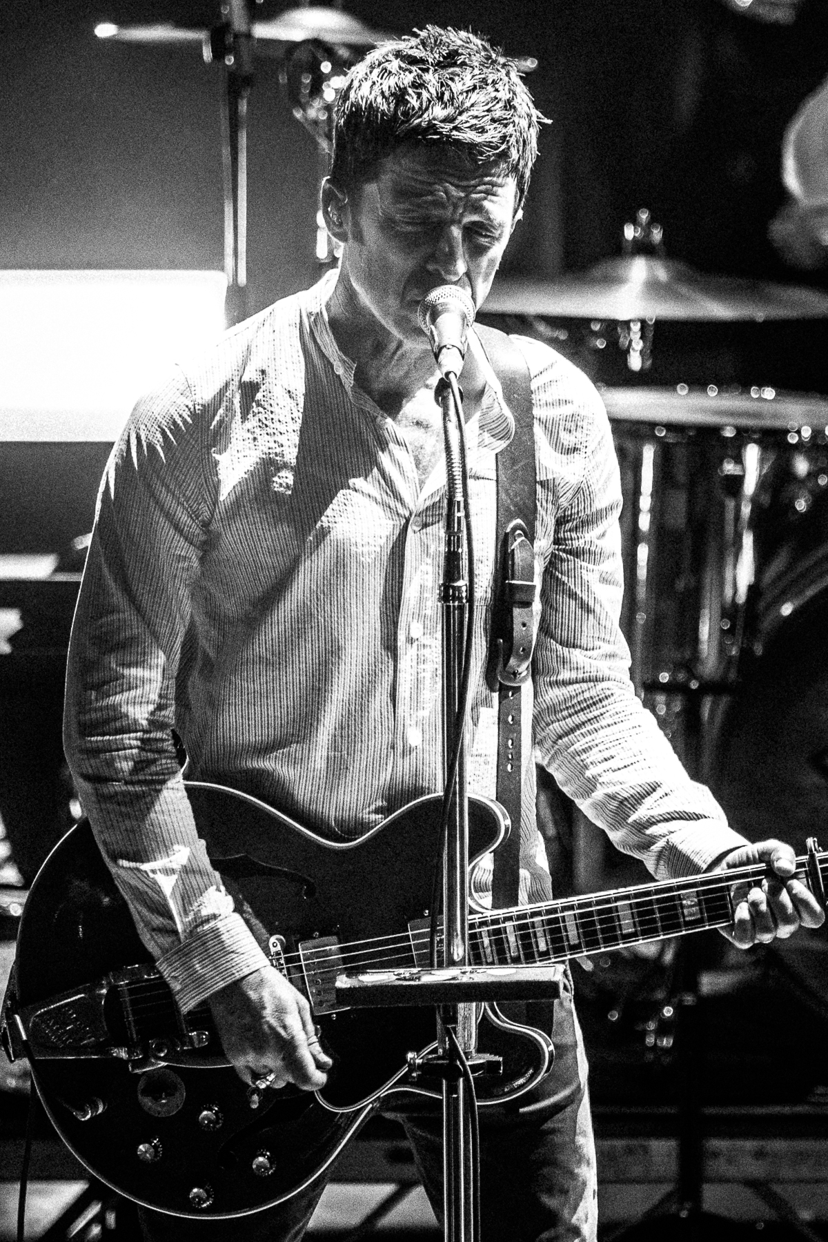 Noel Gallagher sings at a microphone stand with a black semi-hollow guitar, photographed in black and white.