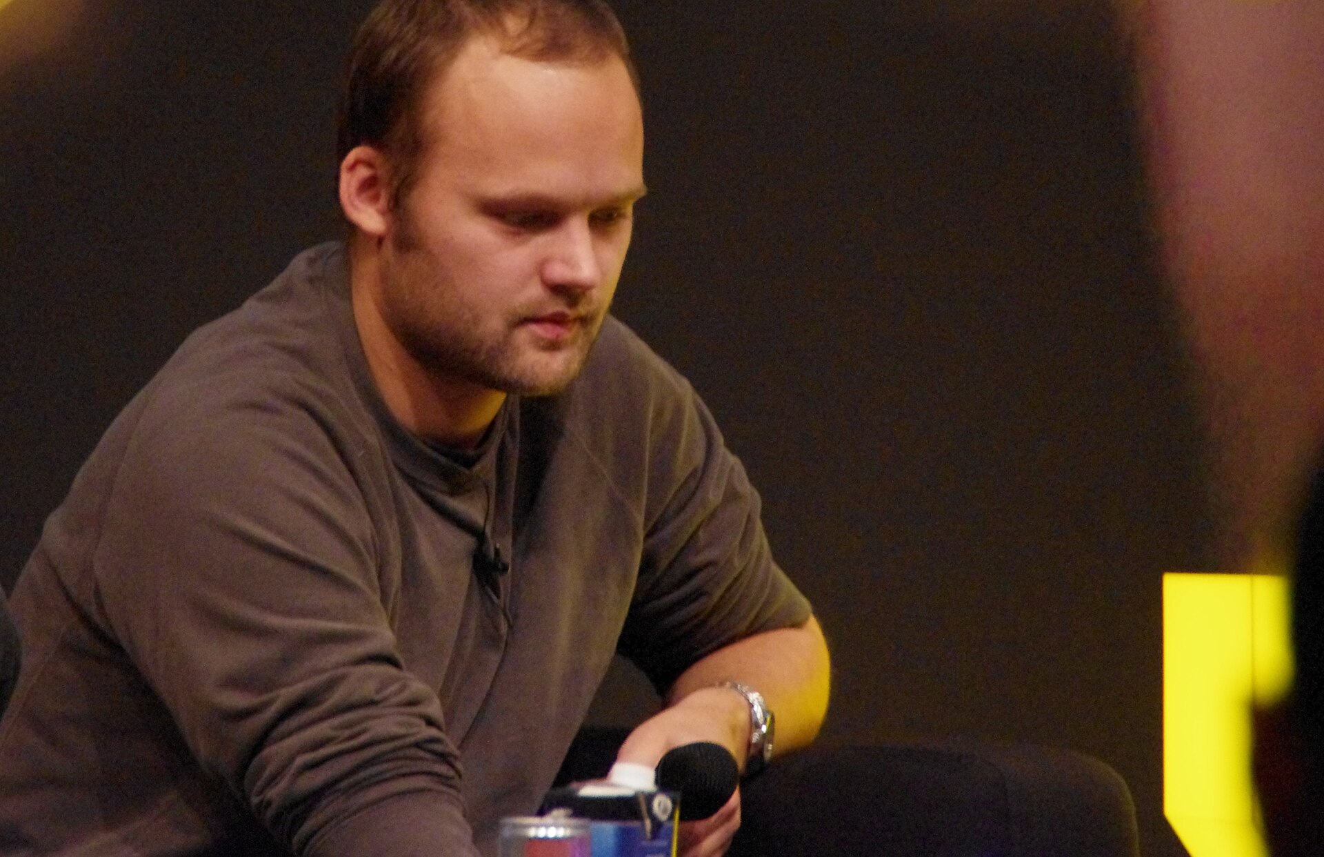 Nik Roos of Noisia sits looking down in a grey long-sleeved shirt, a handheld microphone in front of him, against a dark backdrop.