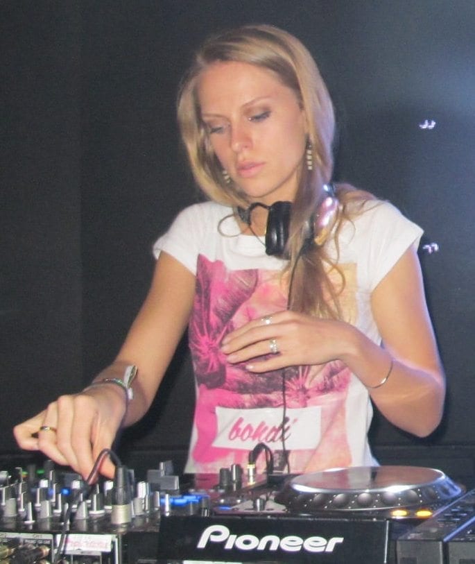 Nora En Pure reaches for a knob on a mixer behind two Pioneer CDJ decks, headphones around her neck.