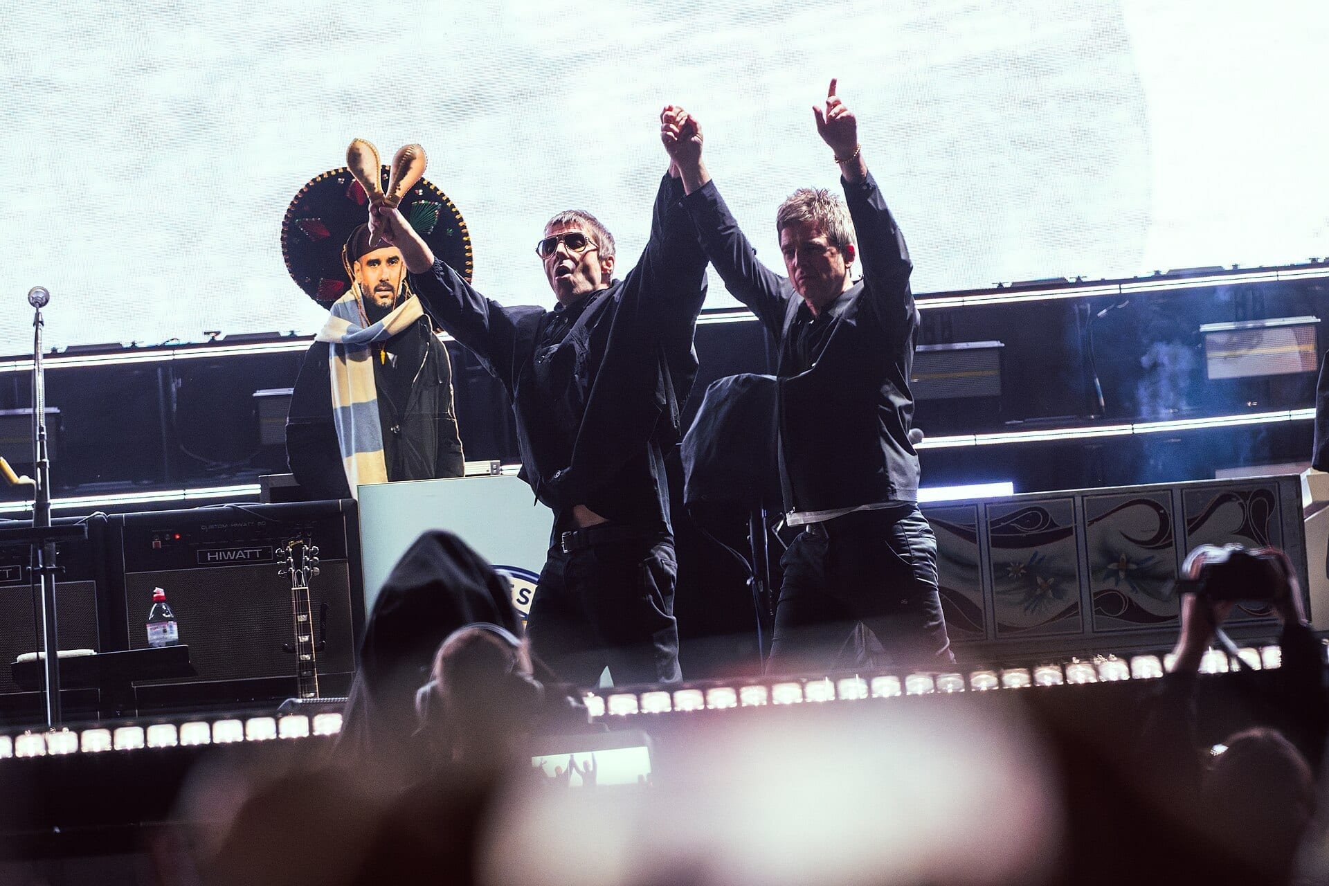Liam and Noel Gallagher stand centre stage with their hands clasped and raised above their heads, both in black, a bandmate in a sombrero holding maracas behind them.