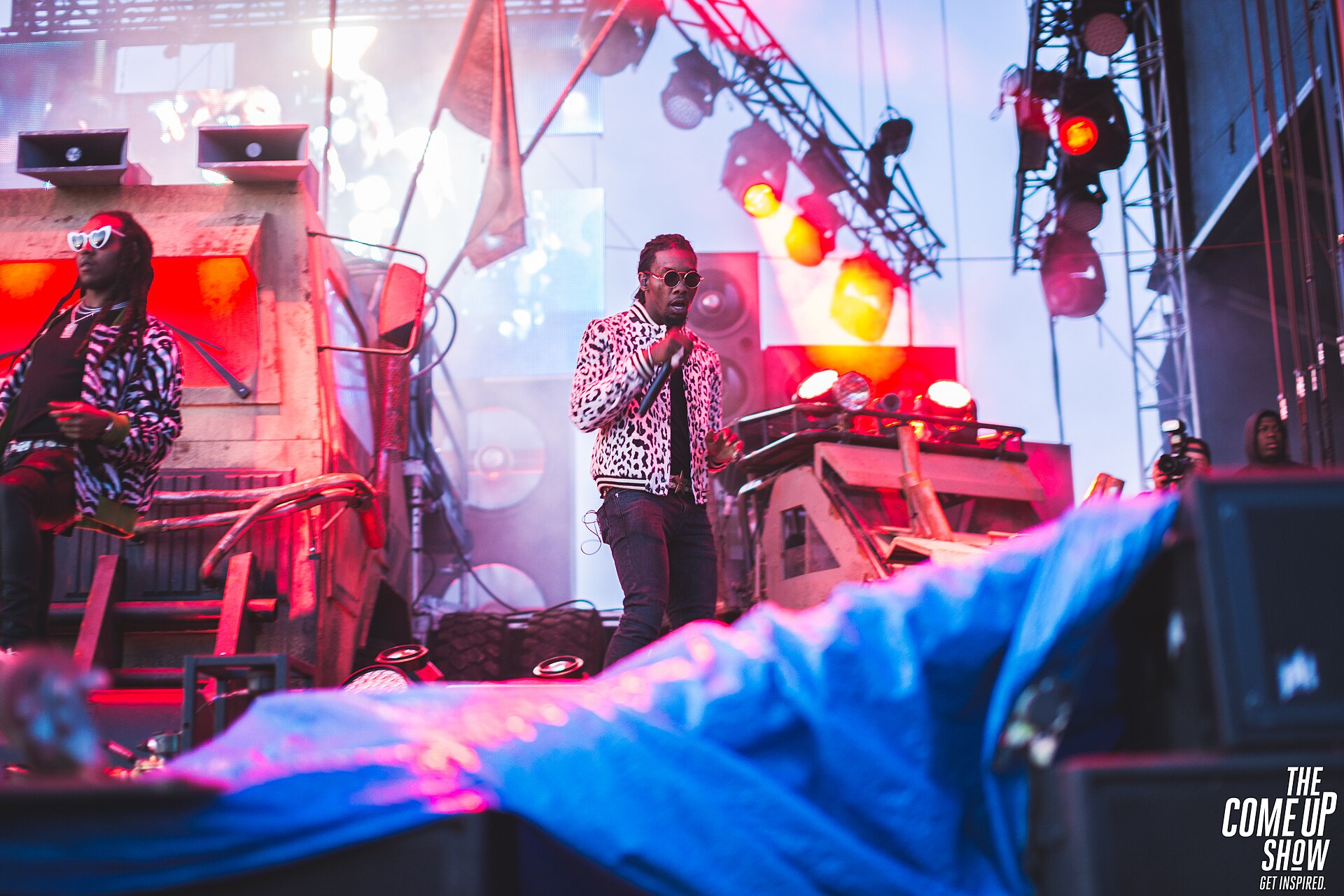 Offset raps into a handheld microphone in a leopard-print jacket and round sunglasses on a festival stage built to look like a wrecked truck, a bandmate at the left.