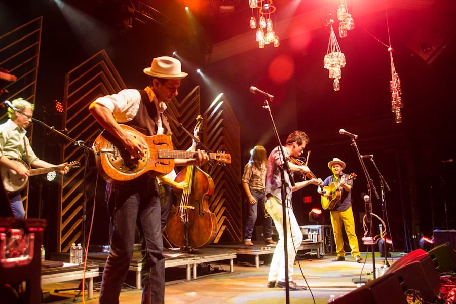 Old Crow Medicine Show onstage in warm light, a player in a straw hat working a resonator guitar in front, a fiddler and an upright bass behind, mason-jar lights hanging overhead.