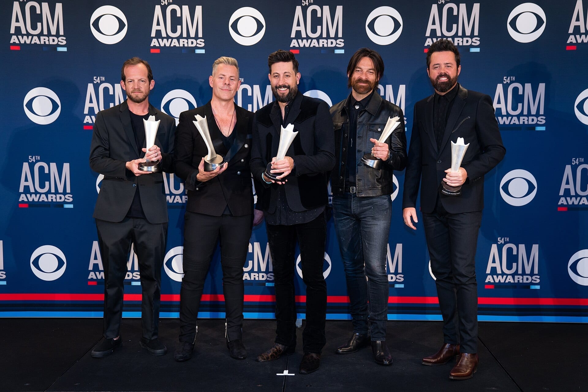 The five members of Old Dominion pose together holding trophies in front of an ACM Awards step-and-repeat backdrop, all dressed in black.