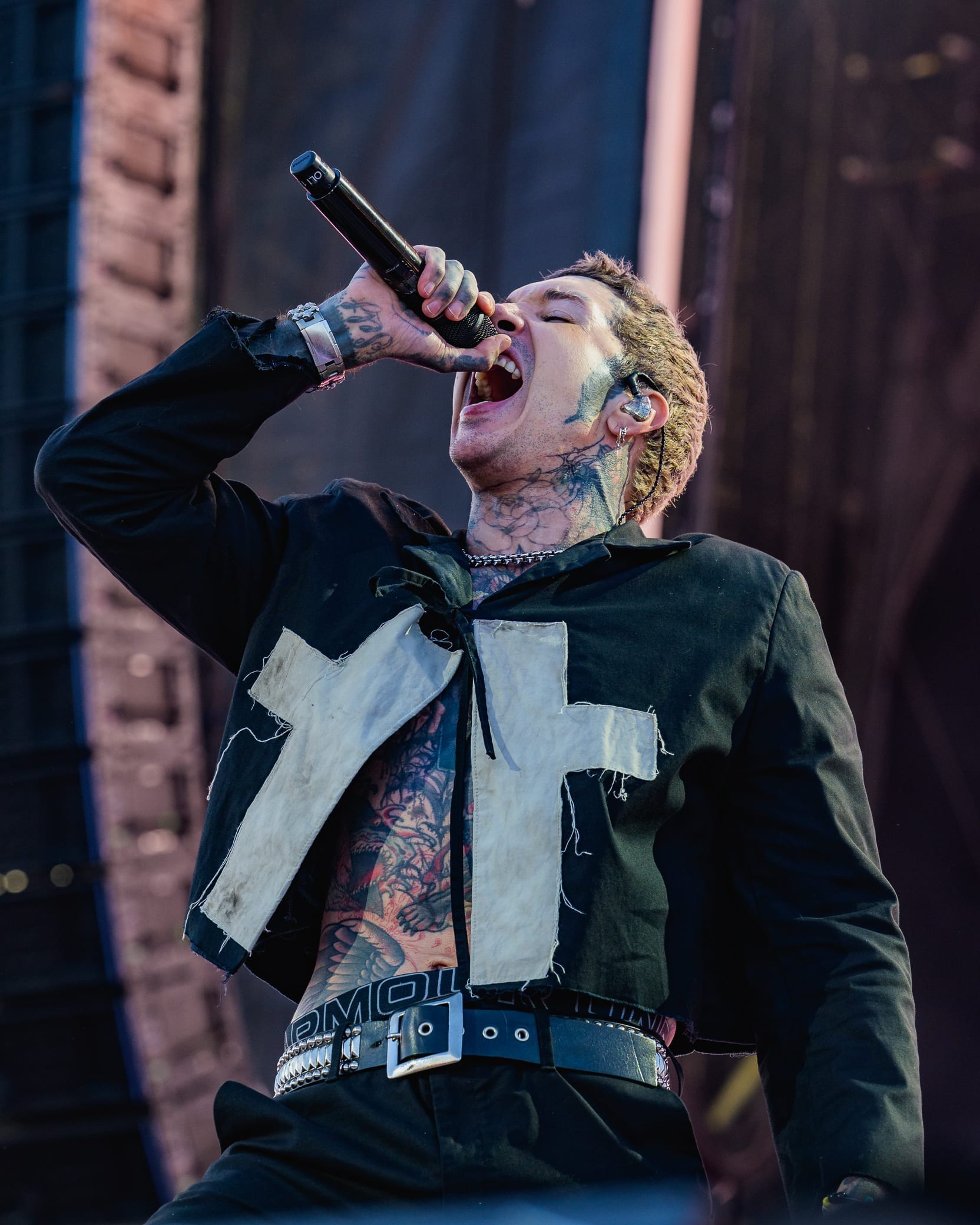Oli Sykes, his face and neck heavily tattooed, sings into a microphone with his head tilted back, wearing a dark jacket with white cross-shaped patches.
