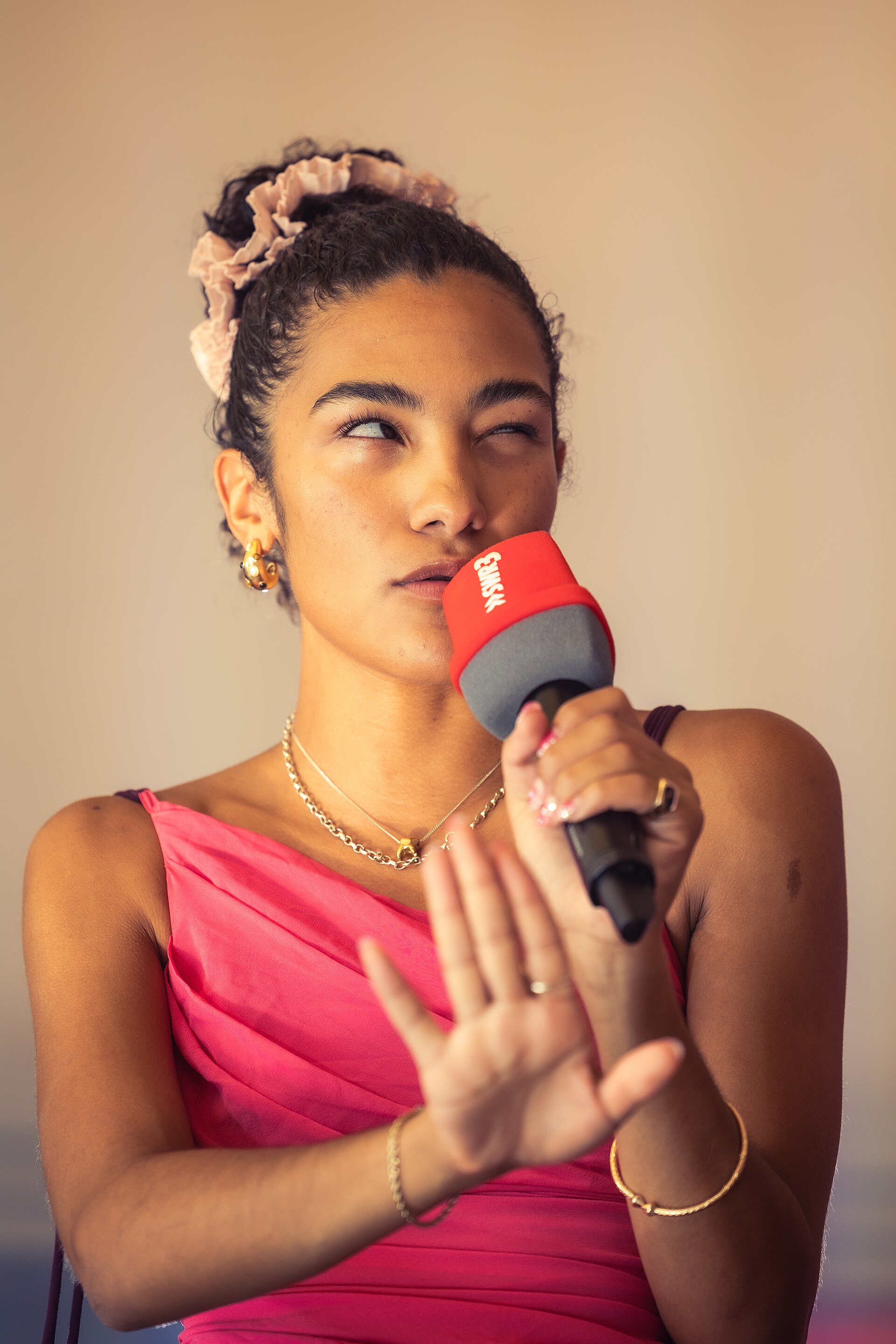Olivia Dean holds a red radio microphone and gestures with one hand, hair up in a pink scrunchie, wearing a pink top and gold hoops.