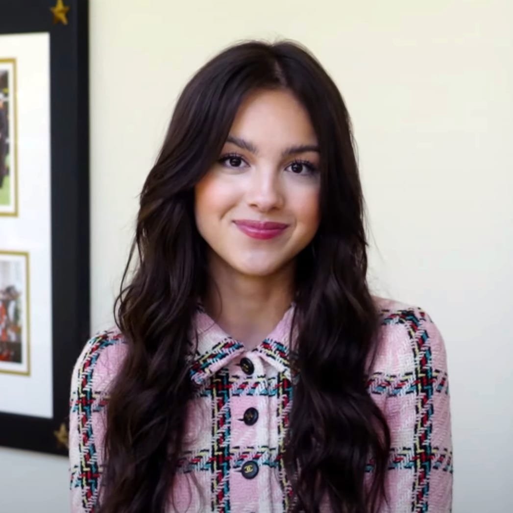 Olivia Rodrigo smiles at the camera, wearing a pink tweed jacket with her long dark hair loose over her shoulders.