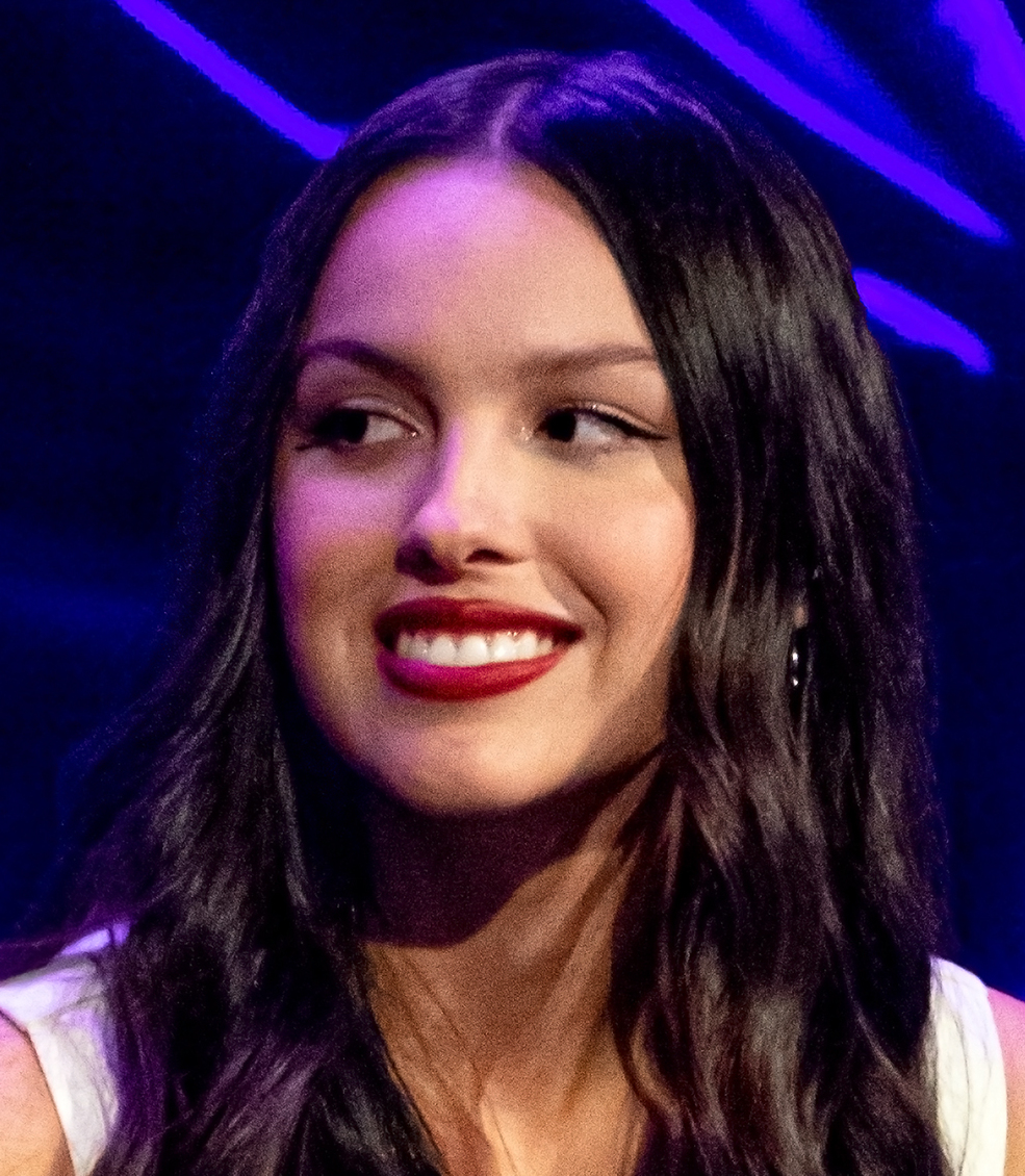 Olivia Rodrigo smiles and looks off camera, dark hair loose over a white top, lit purple and pink.