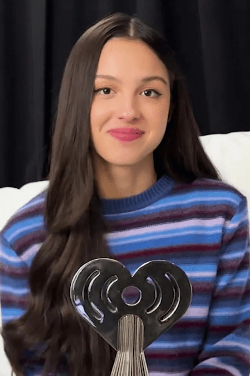 Olivia Rodrigo smiles slightly while holding a heart-shaped iHeartRadio trophy, wearing a striped sweater in front of a dark curtain backdrop.