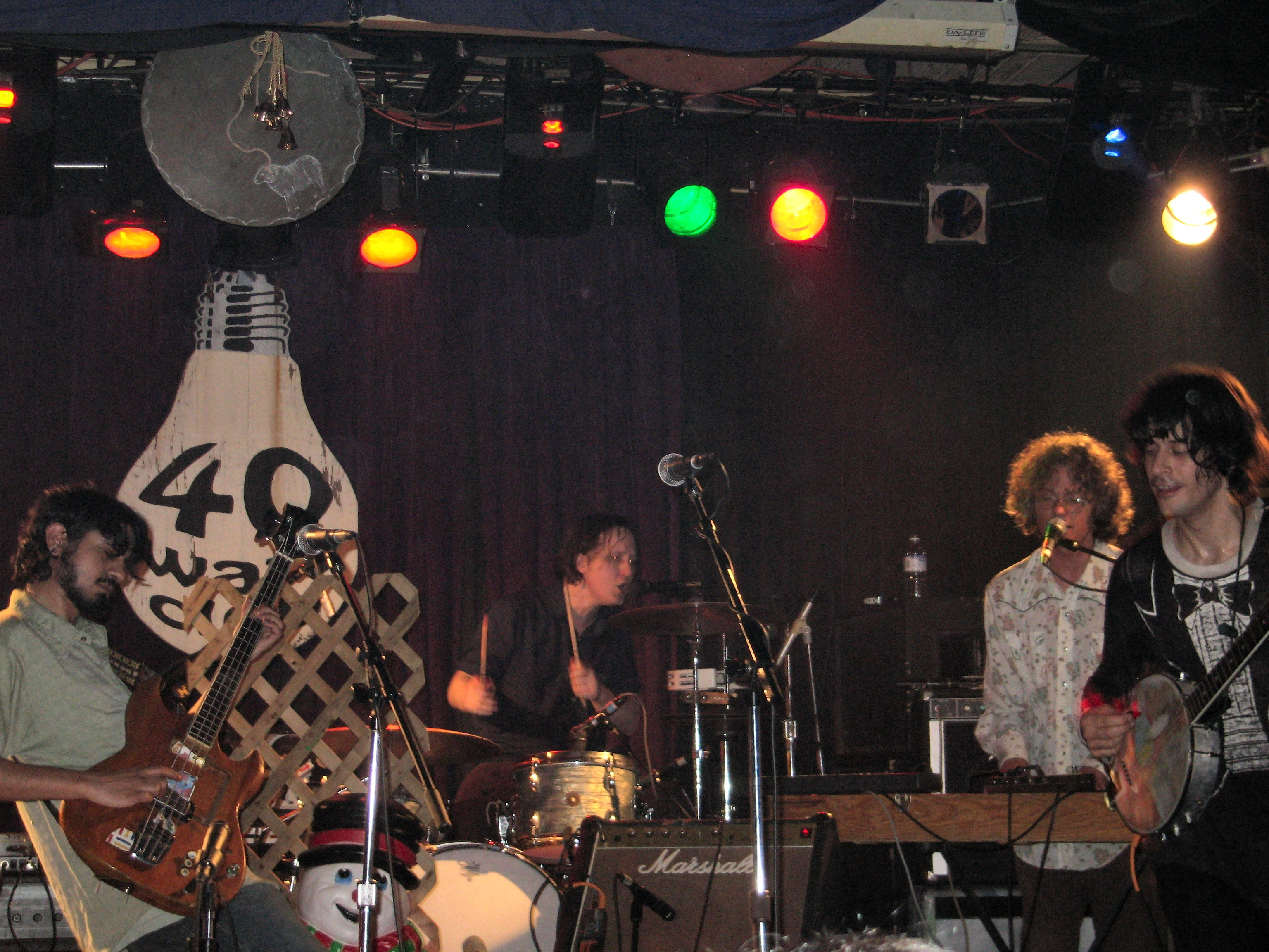 The Olivia Tremor Control play a small club stage under a painted light-bulb backdrop reading 40 Watt, a bassist at the left and a drummer in the middle.