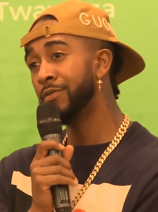Omarion, wearing a tan cap and a gold chain over a dark t-shirt, speaks into a handheld microphone in front of a green backdrop.