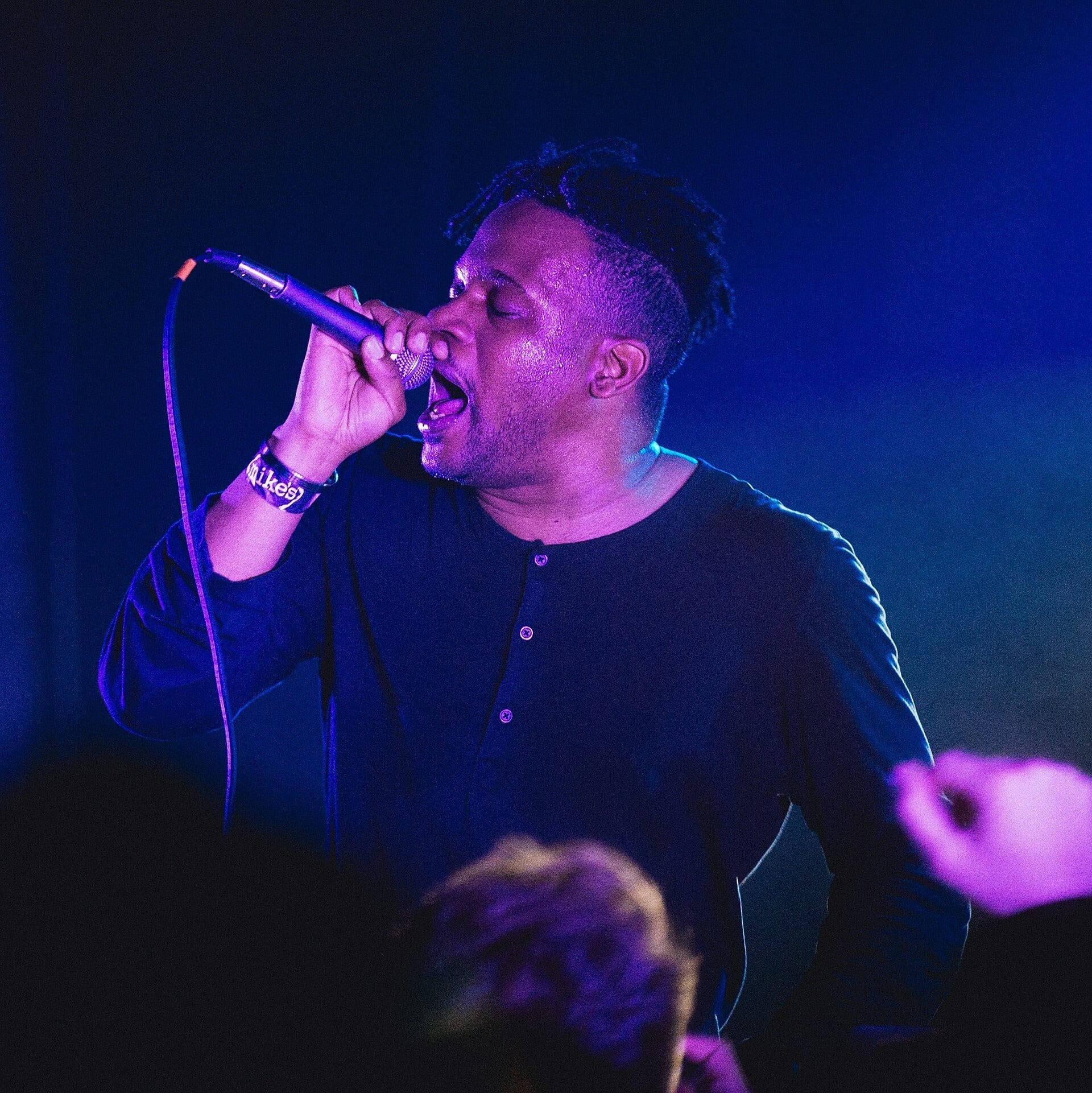 Open Mike Eagle sings into a microphone held close to his mouth under blue and purple stage light, wearing a dark button-up shirt and a wristband reading mike's.