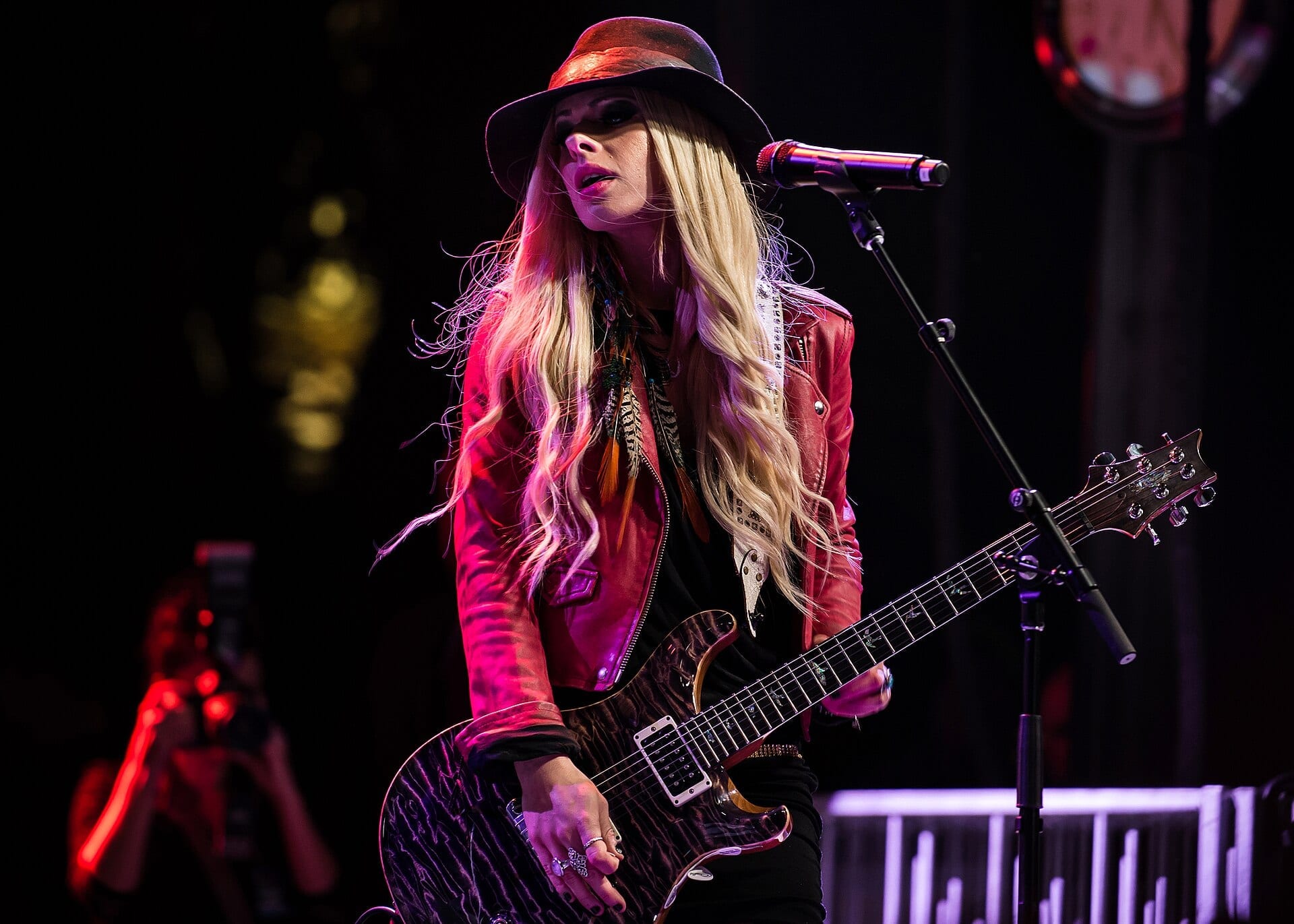 Orianthi plays a figured-top electric guitar at a microphone stand in a black hat and pink leather jacket, long blond hair over her shoulders under magenta stage light.
