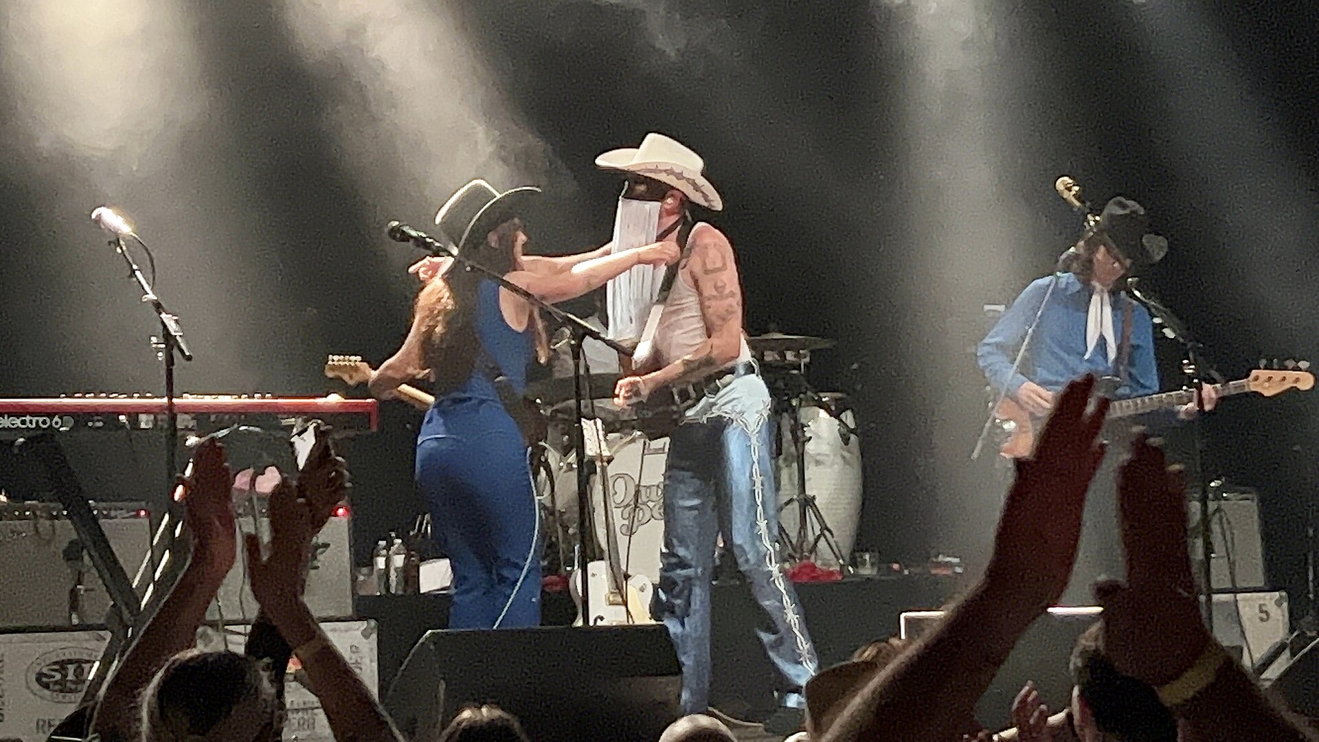 Orville Peck, in his fringed mask, white cowboy hat and cut-off jeans, leans toward a bandmate at a microphone while the crowd raises their hands in front of the stage.