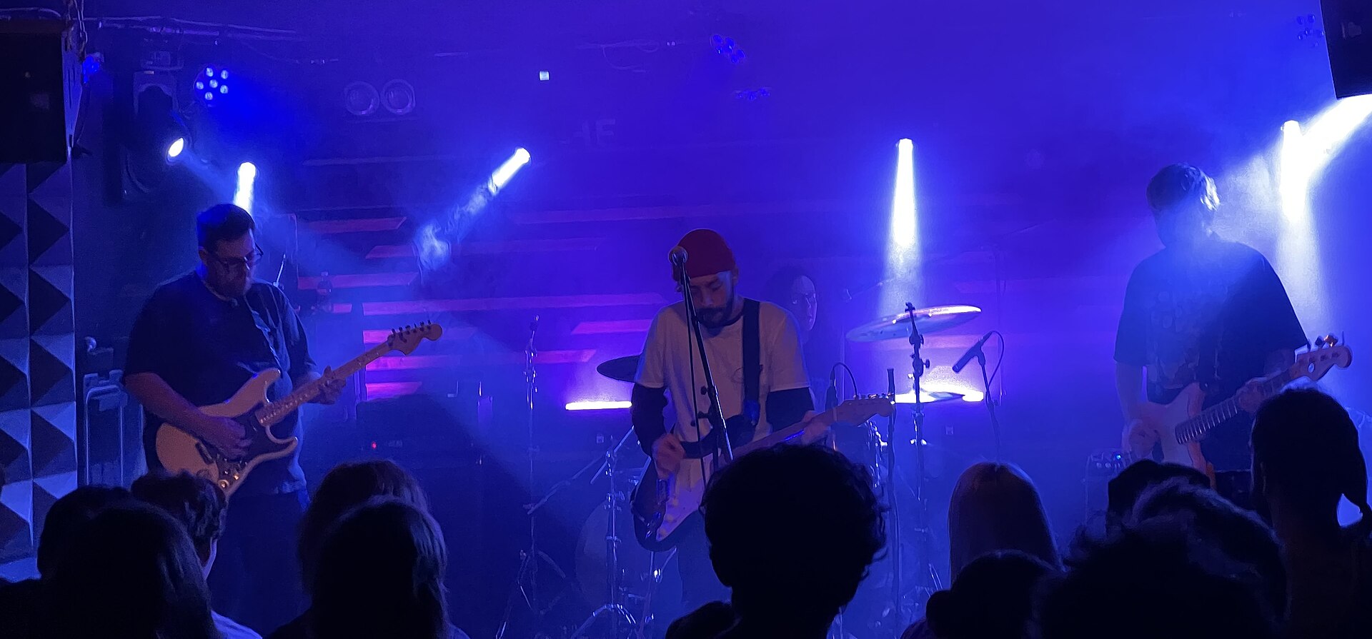 Oso Oso on a small blue-lit club stage, the singer at a centre microphone with guitarists either side.